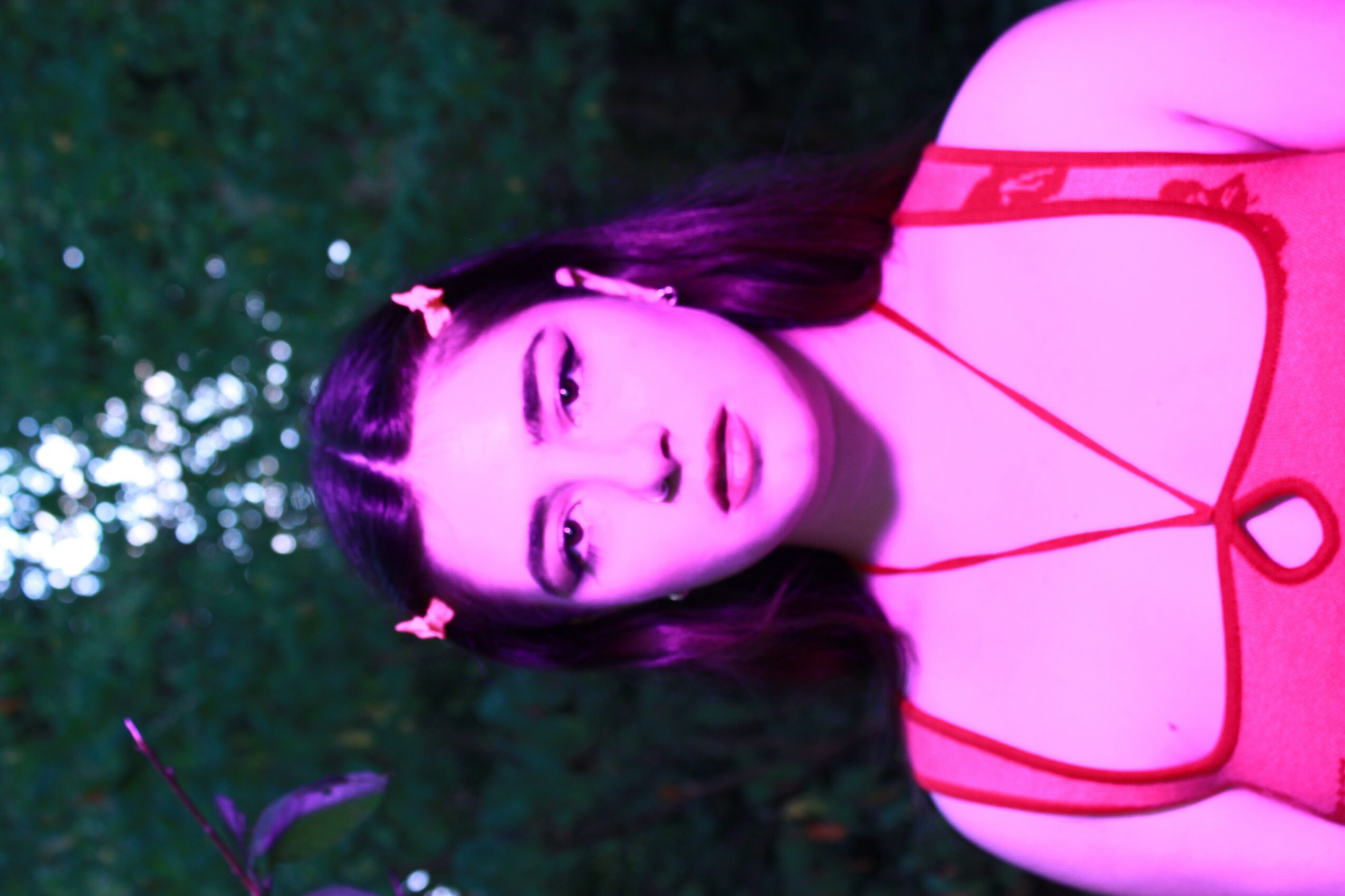Young woman with long dark hair and makeup, wearing a pink top with red straps, outdoors at night with trees in background.