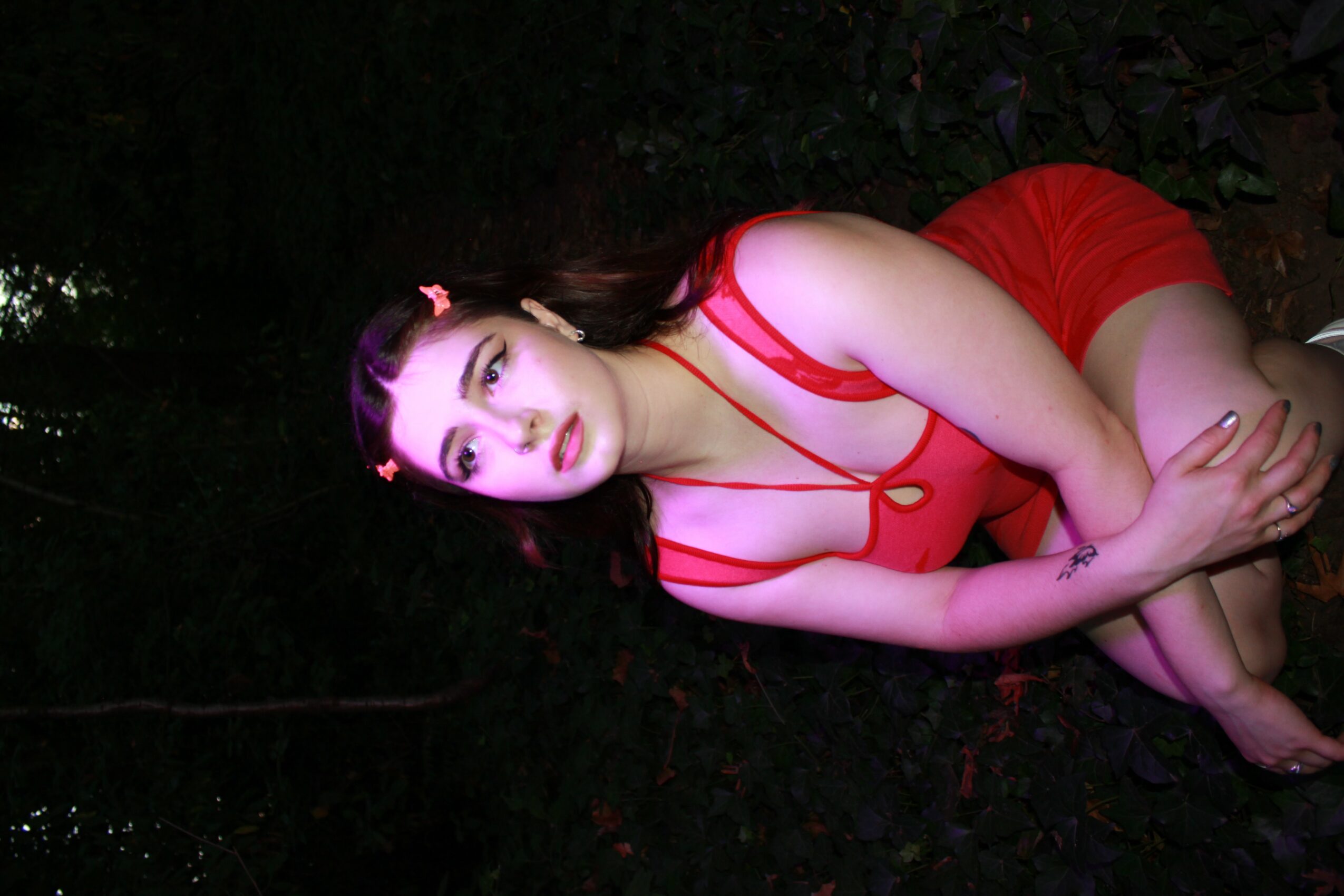 Young woman in red outfit lying on dark ground, looking at camera with makeup and pink hair accessories.