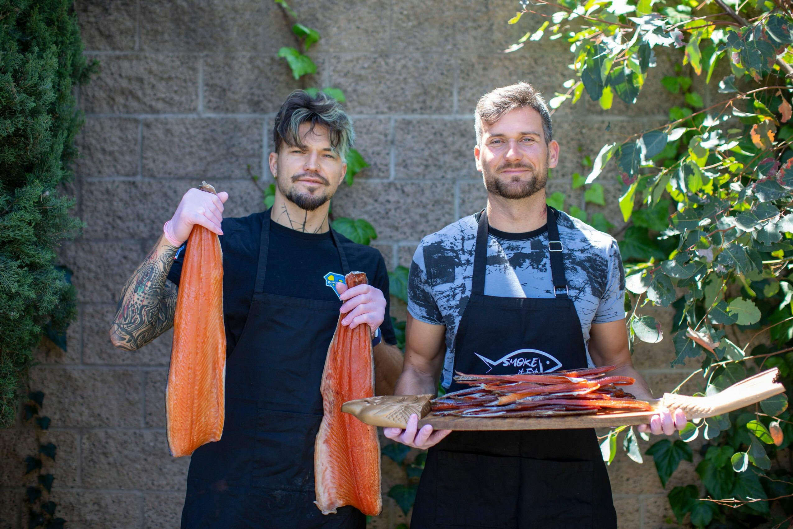 Two men stand outdoors holding a tray of food, surrounded by greenery and a stone wall.