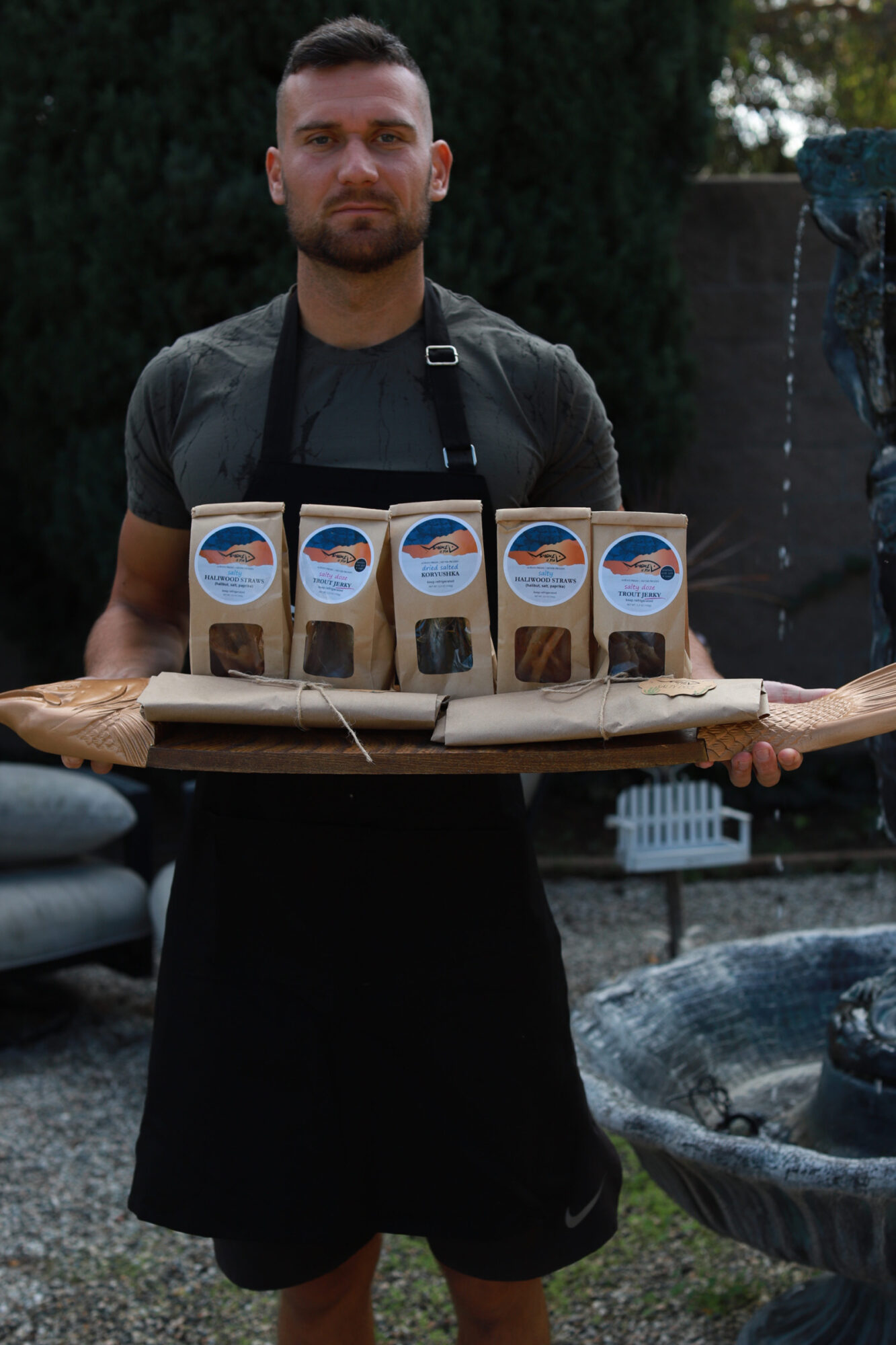 Person standing outdoors with a table of six coffee cups, trees in the background, and a person sitting nearby.