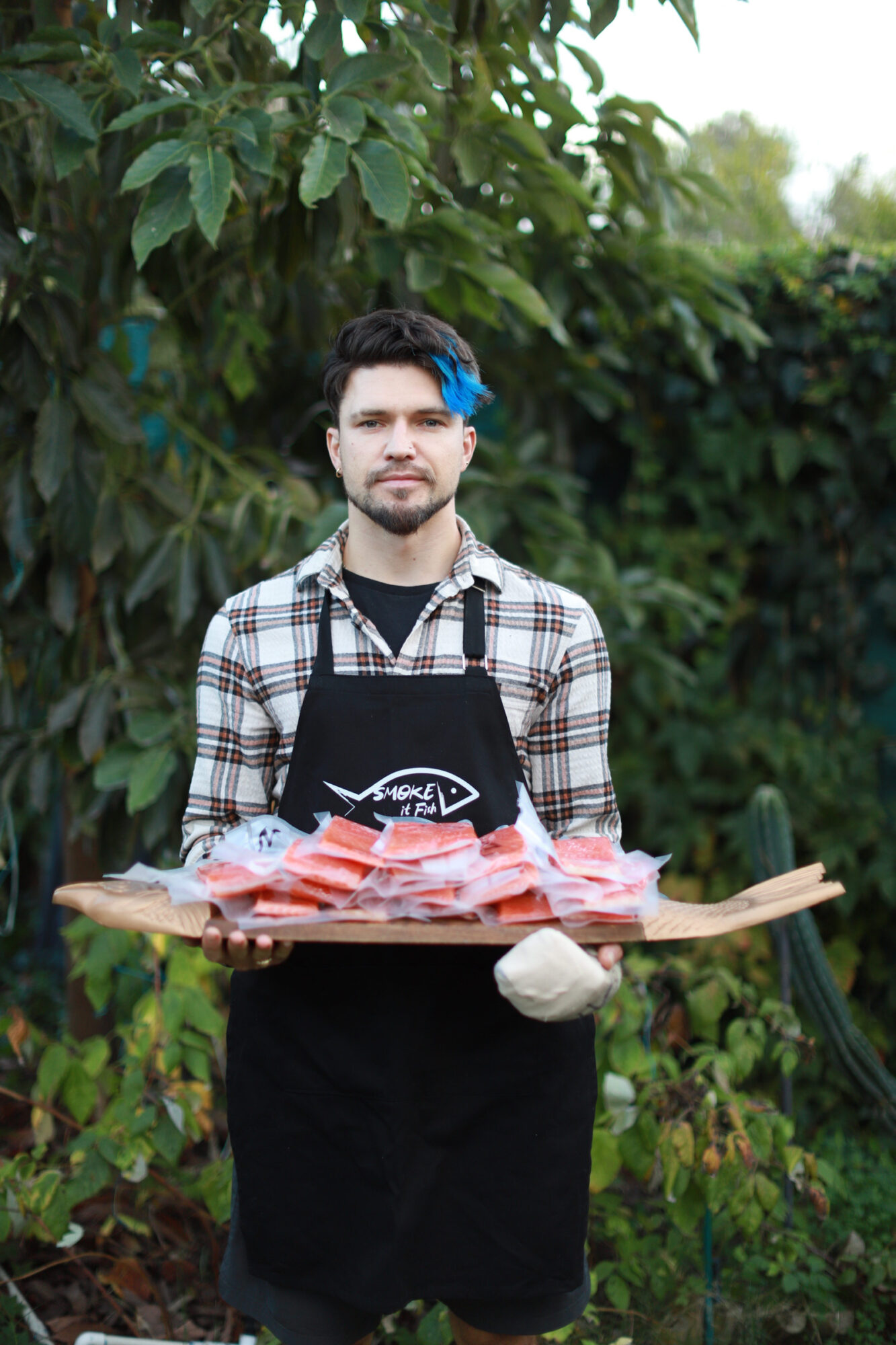 Person with short dark hair and makeup, wearing a plaid shirt over a black graphic t-shirt, standing outdoors with greenery.