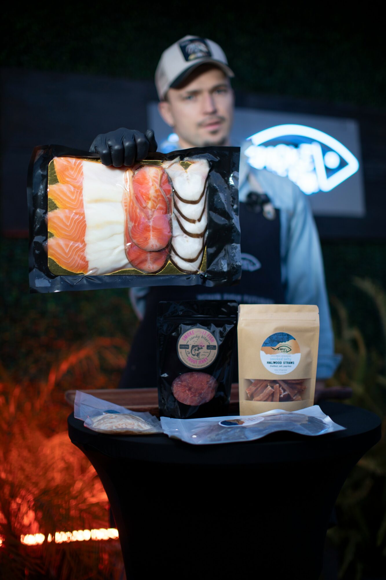 Person holding a tray of sushi, with food items and packaging on a table, background dark with a logo. Word count: 20