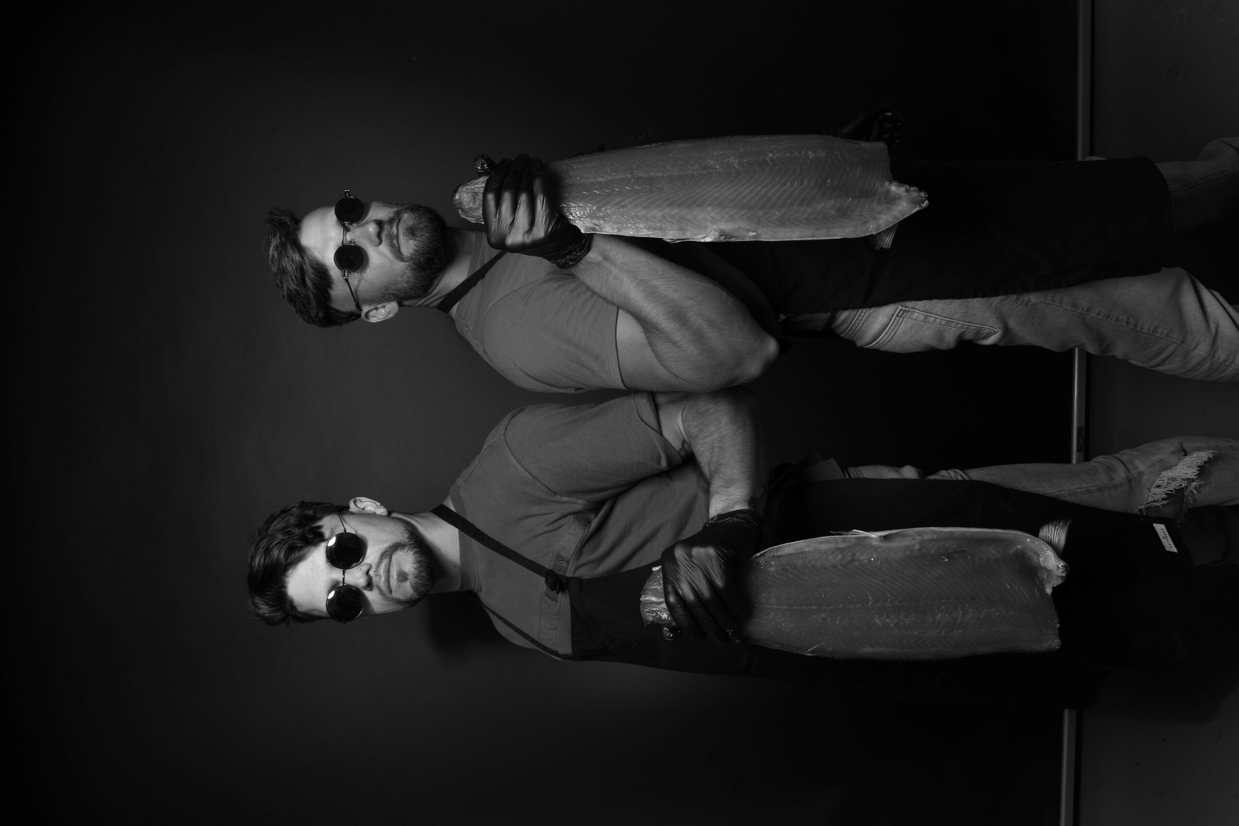 Two people wearing sunglasses and jackets, standing against a dark background, one holding a drink, both facing sideways.