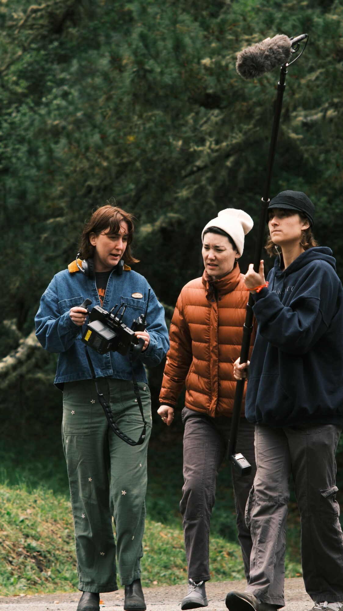 Three people standing outdoors, one holding a camera, engaged in conversation, with trees in the background.