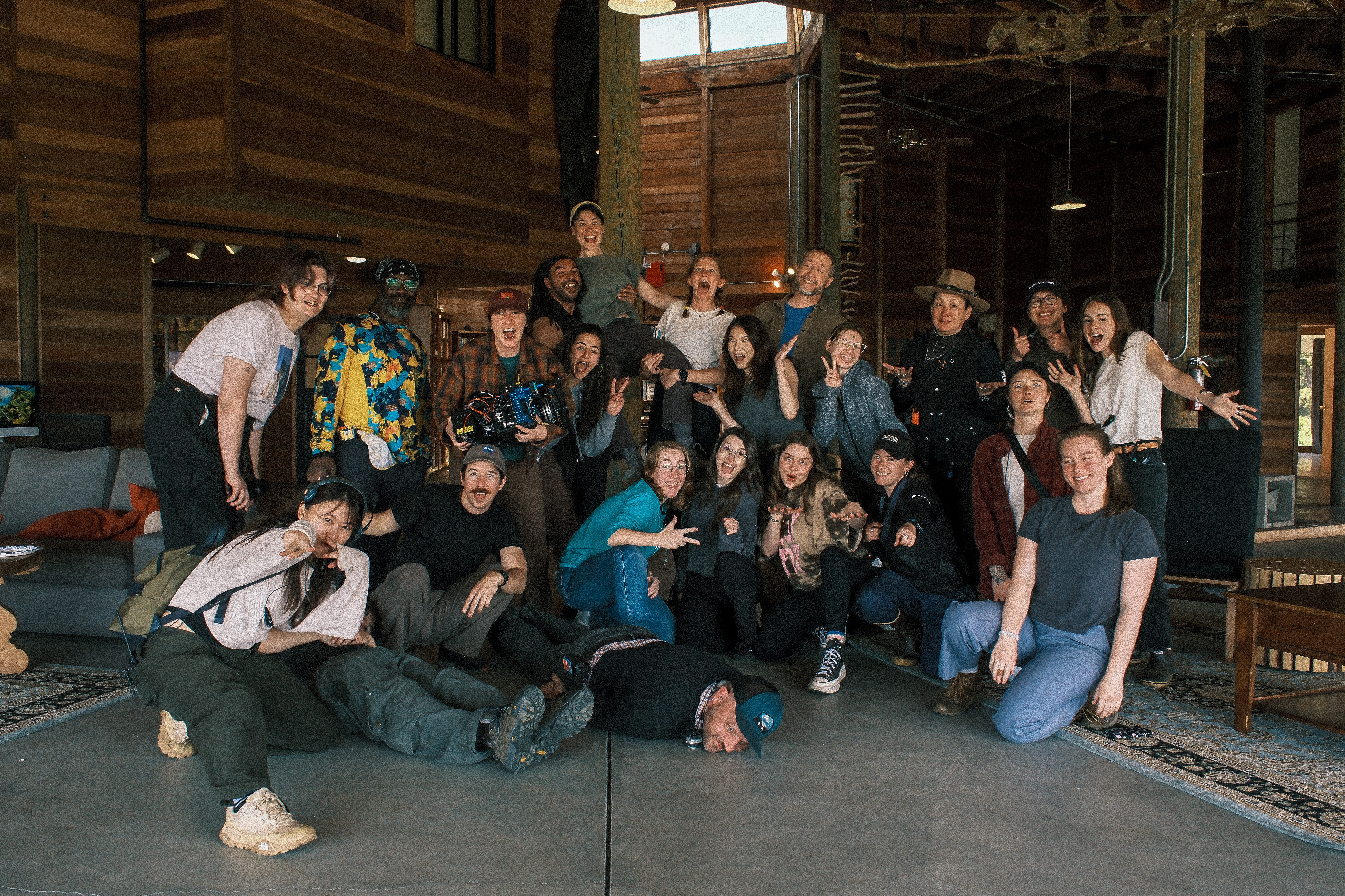 Group of people posing indoors, some sitting and some standing, in a rustic setting with wooden walls and large windows.