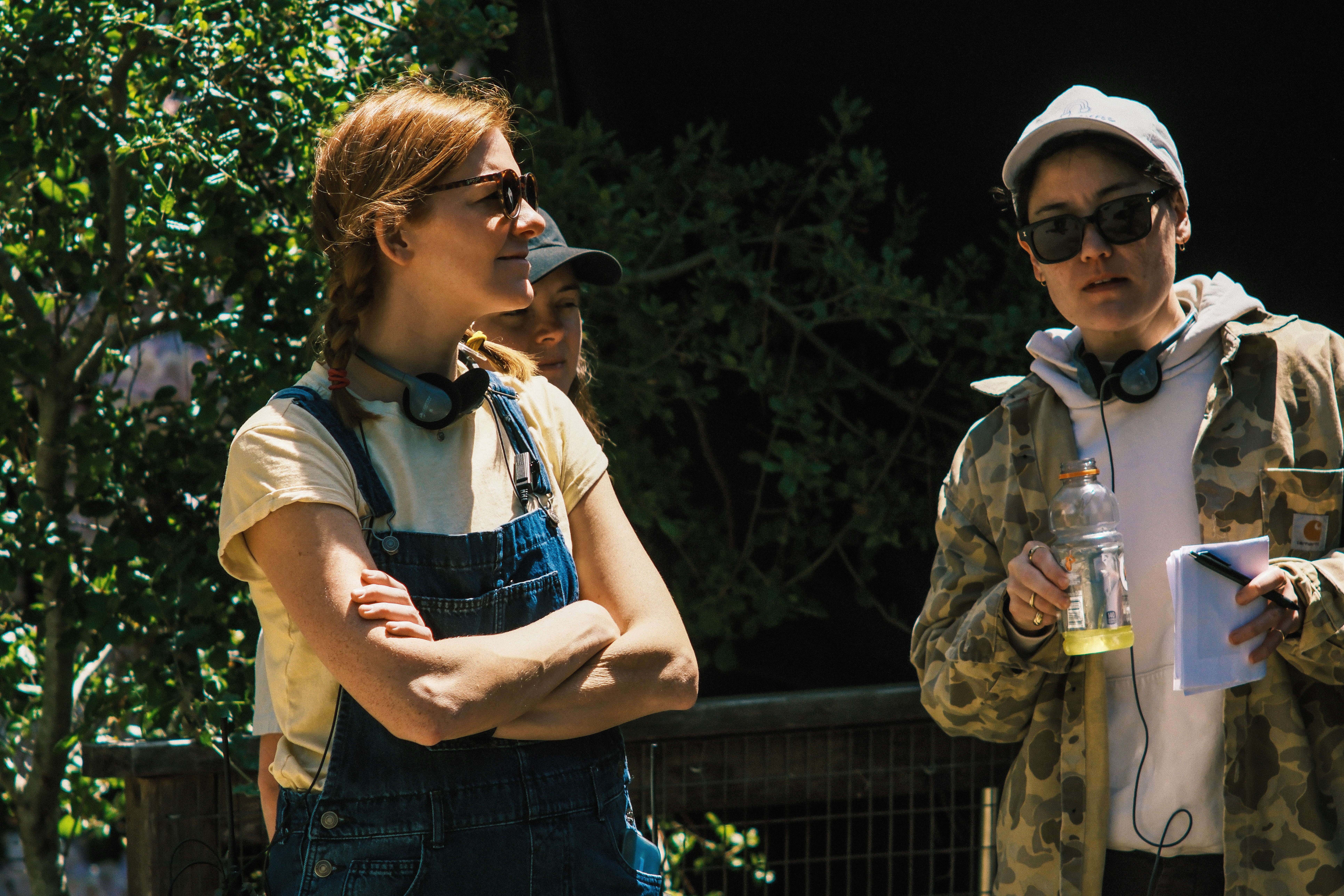 Two women standing outdoors, one with arms crossed, wearing sunglasses and casual clothing, the other holding a drink and a notebook, both with headphones around their necks.