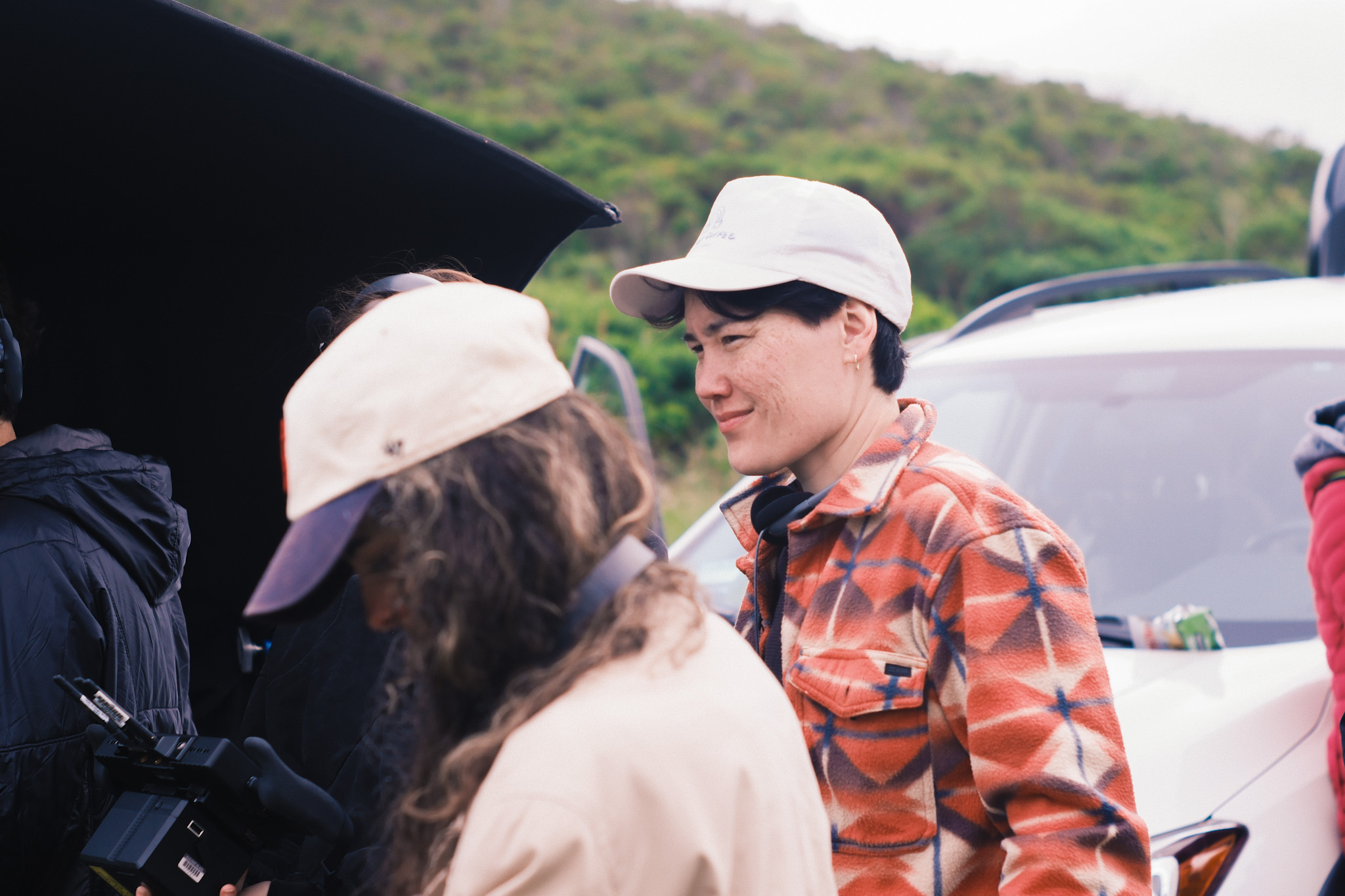 Two people wearing white caps and a woman with long hair are outdoors, smiling and talking, with a car and greenery in background.