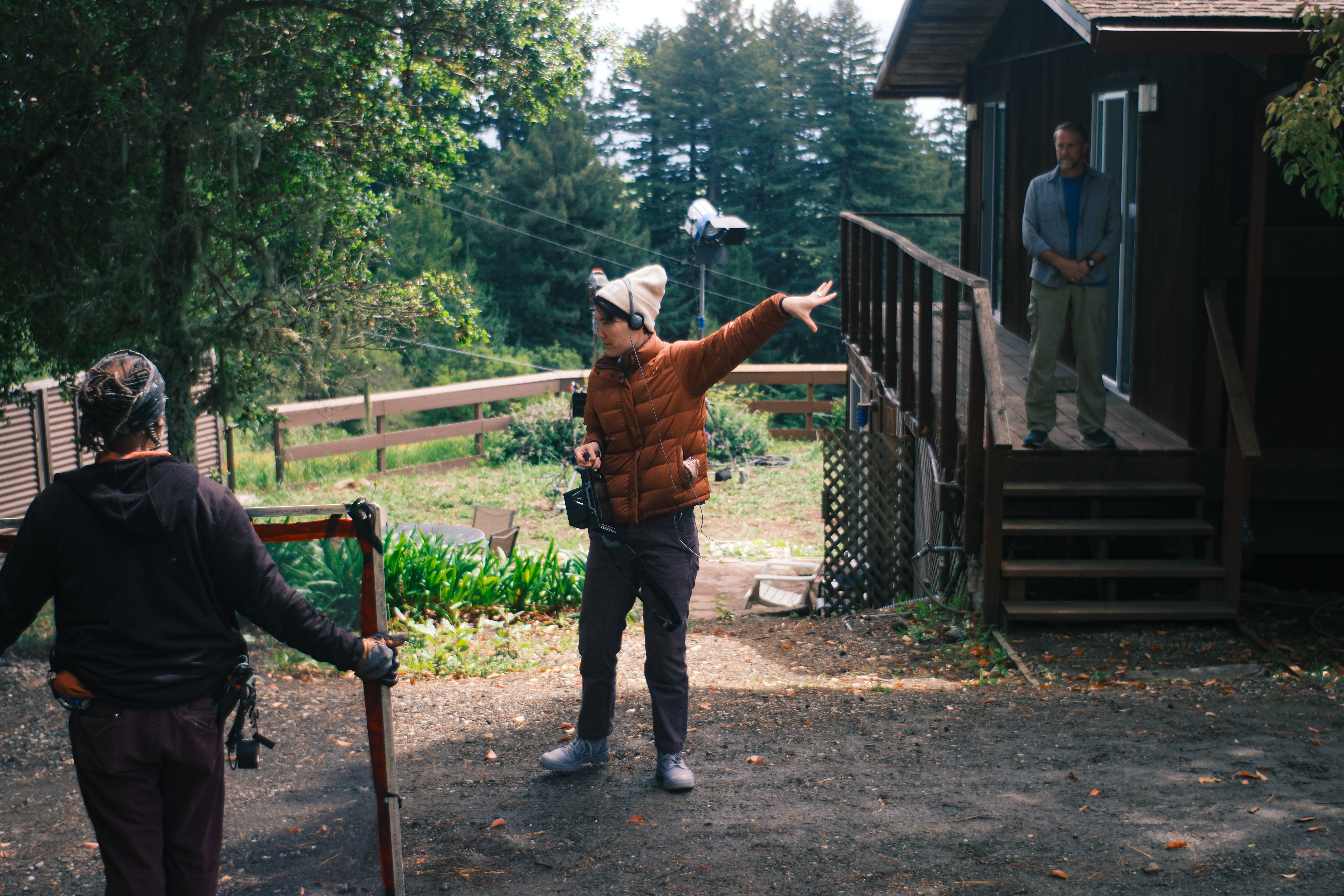 Three people outdoors near a wooden house, one person gesturing with arms outstretched, another holding a cane, and a third on stairs.
