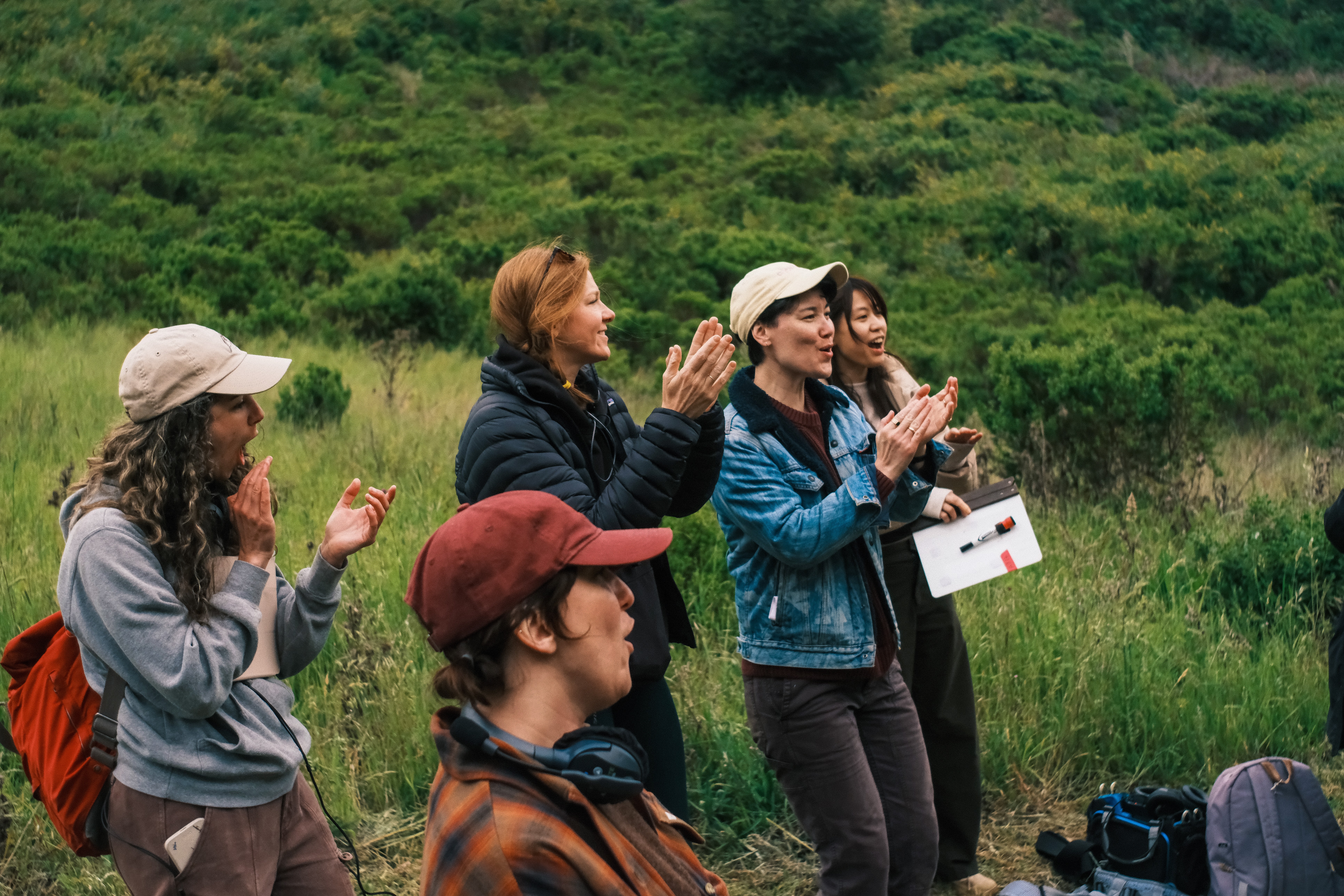 Group of four women and one man standing outdoors in a grassy field, clapping and smiling, with backpacks and camera nearby.