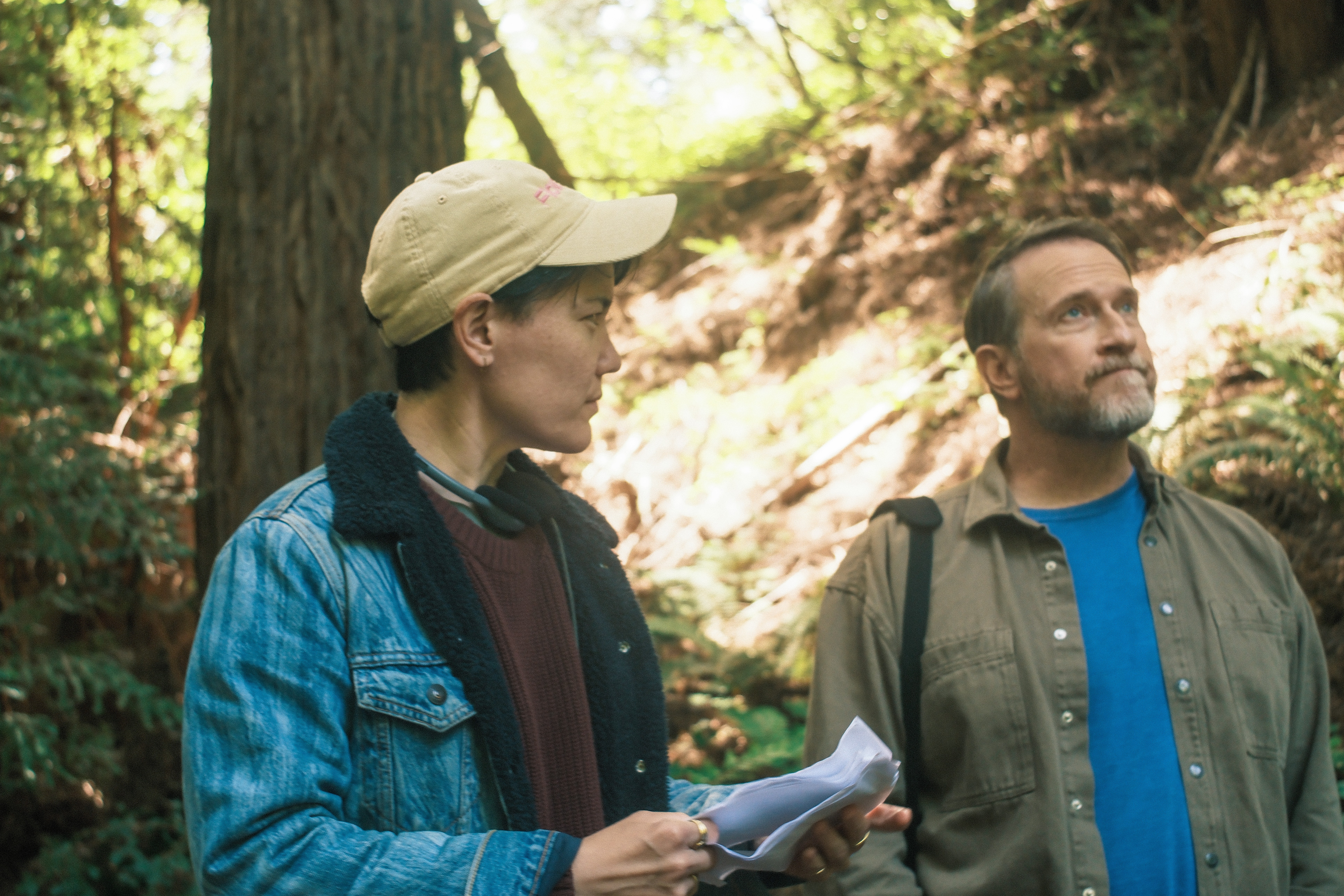 Two people standing outdoors in a forest, one holding papers and the other looking at them.