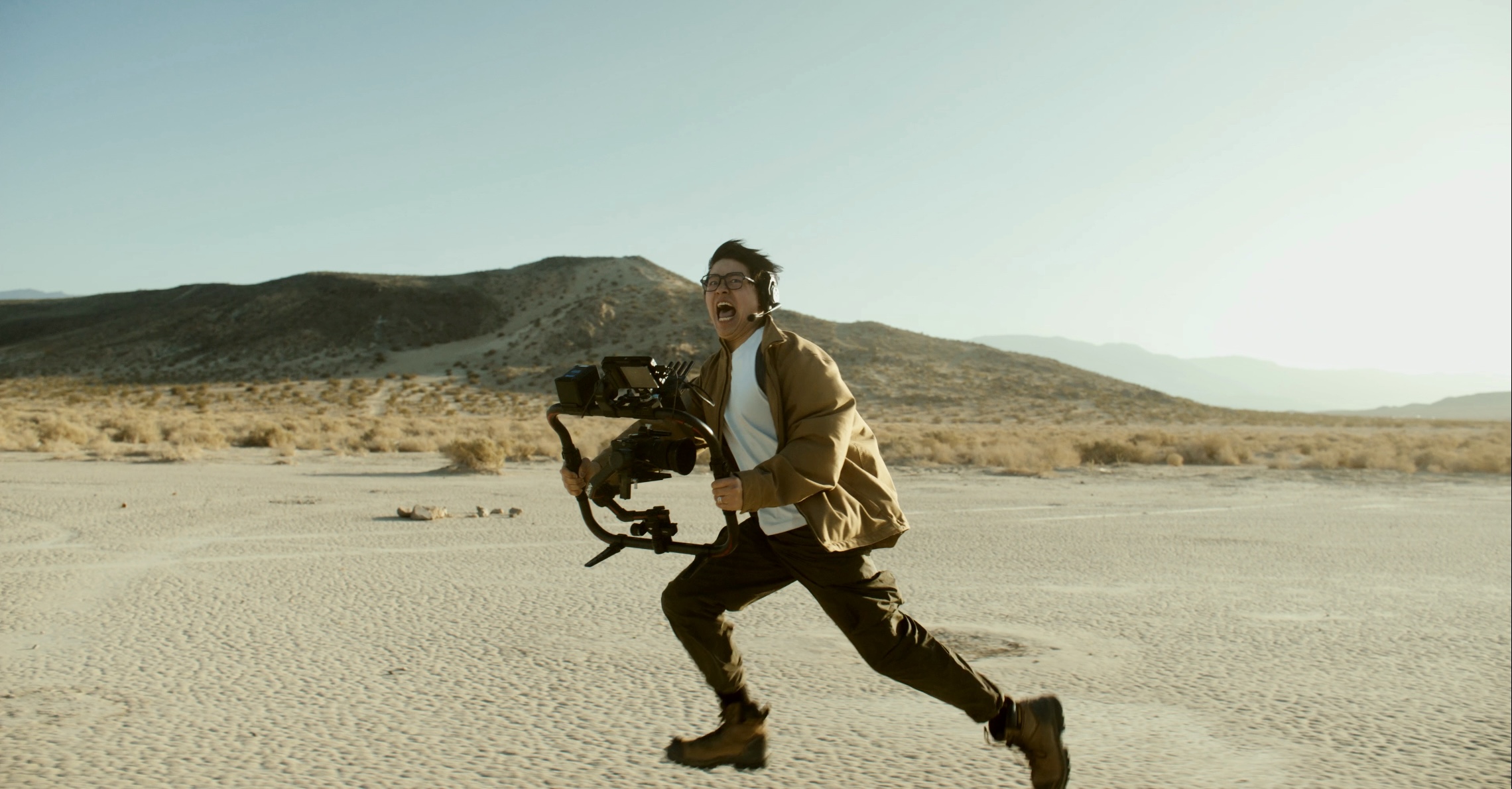 Person running in a desert landscape with mountains in the background, carrying a camera or equipment, wearing outdoor clothing.