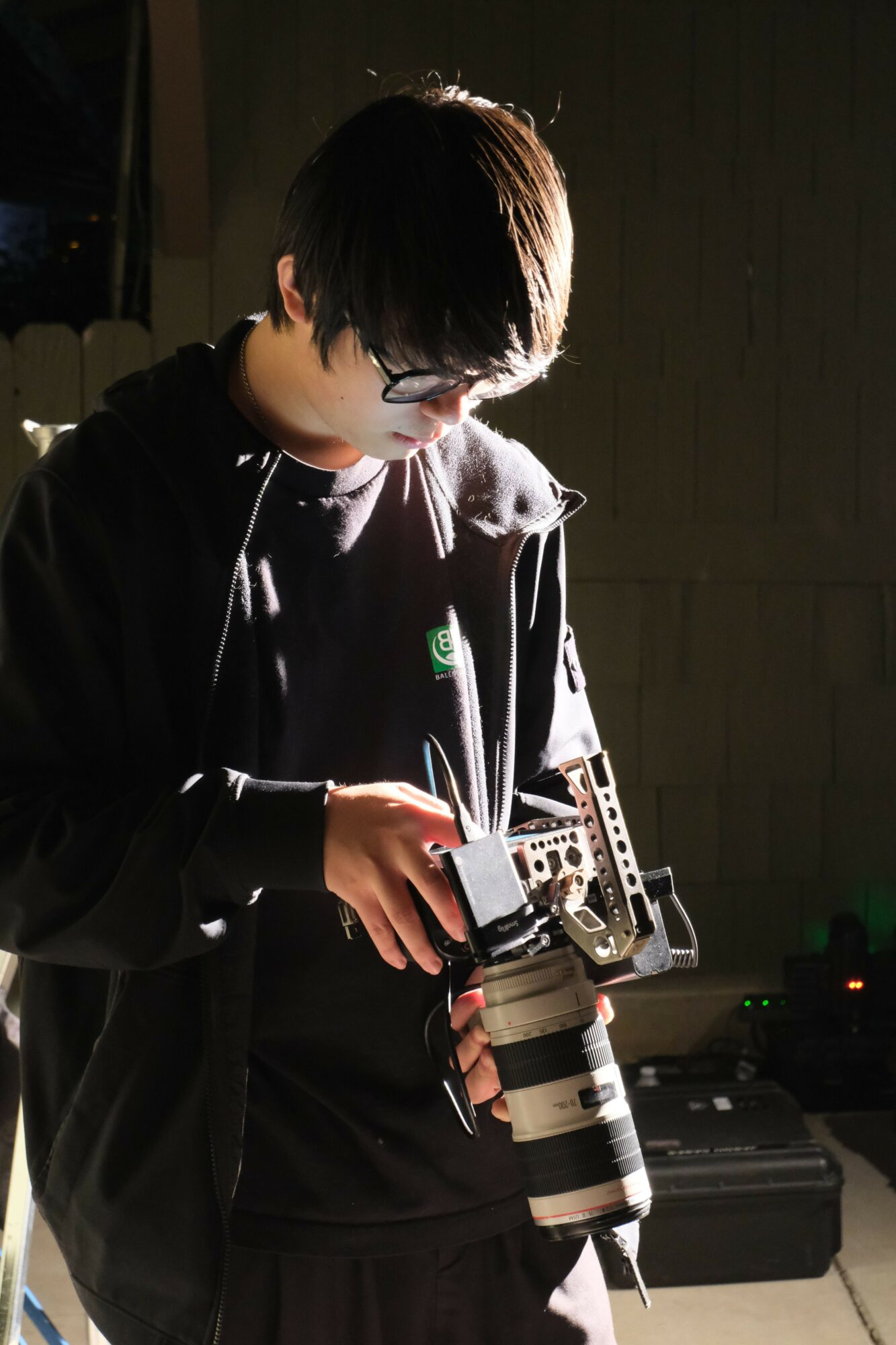 Person with dark hair and glasses holding a camera, standing indoors with equipment on a table behind them.