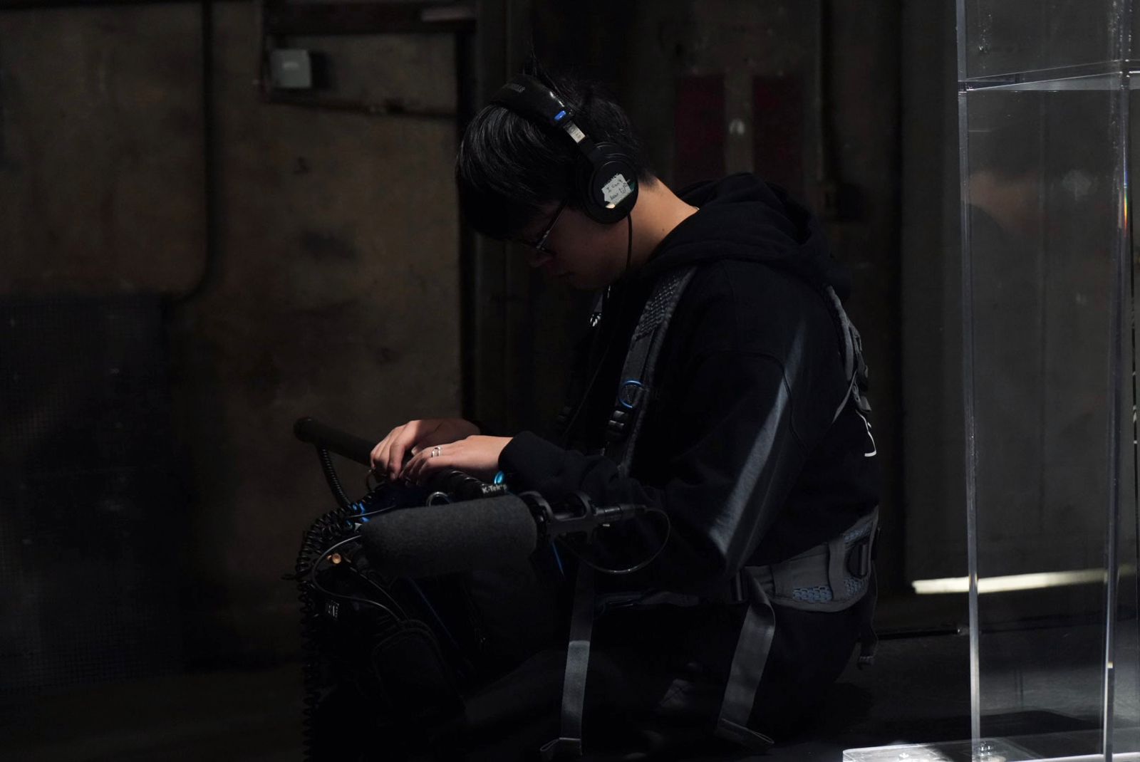 Person sitting on a bench using a smartphone, wearing headphones and a black hoodie, in a dimly lit environment.