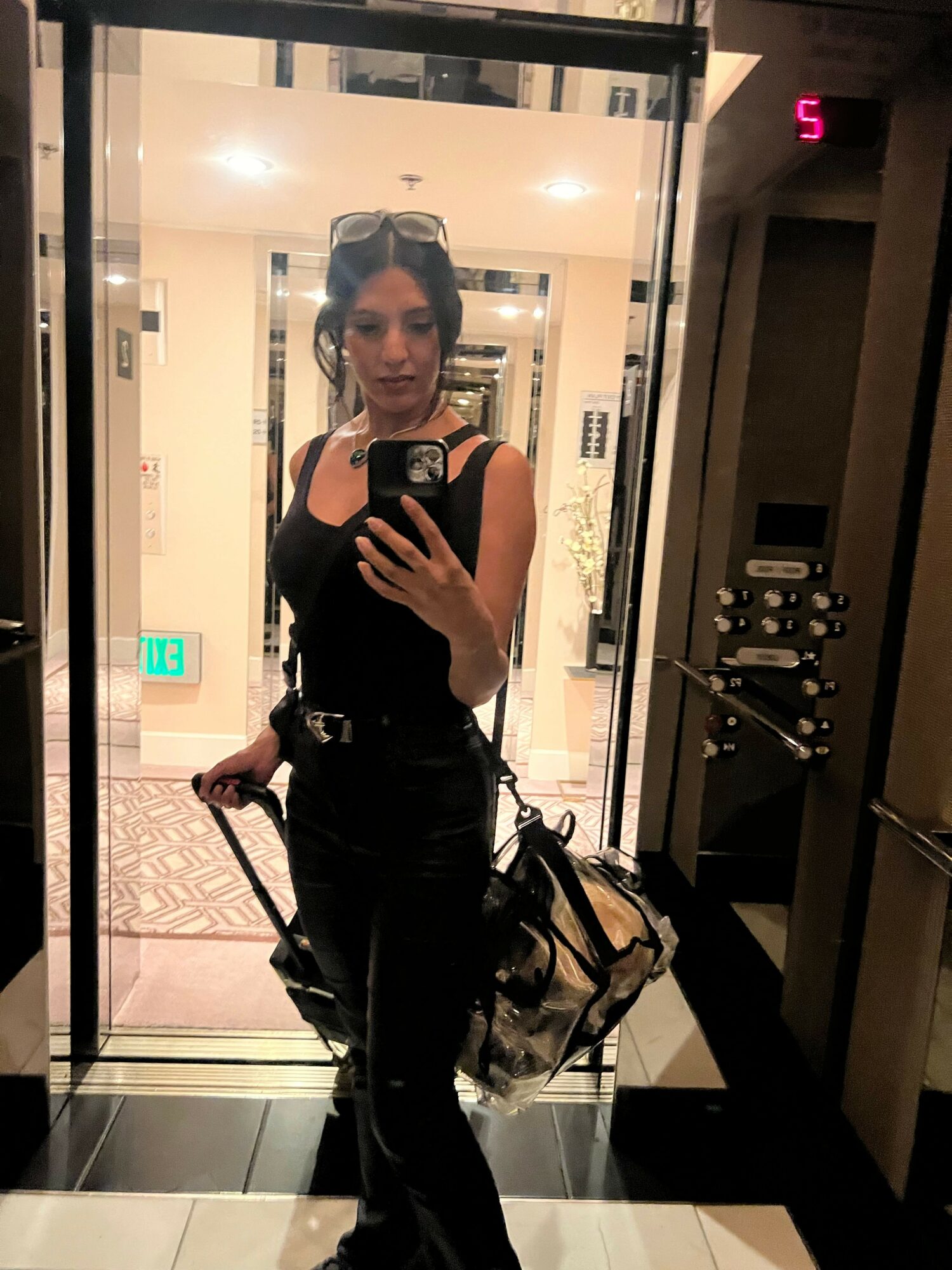 Person taking a mirror selfie in an elevator, holding a bag, with elevator control panel visible behind.