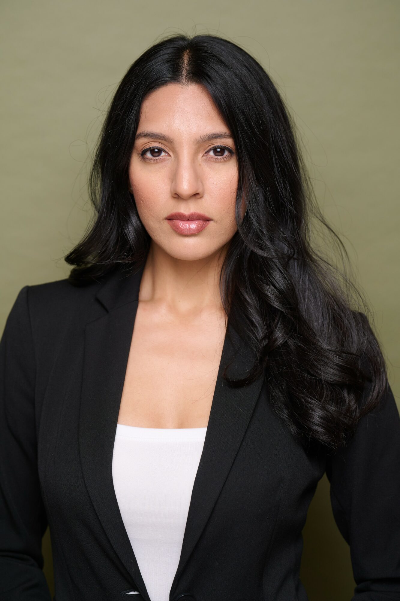 Woman with long dark hair wearing a black blazer and white top, facing forward against a plain background.