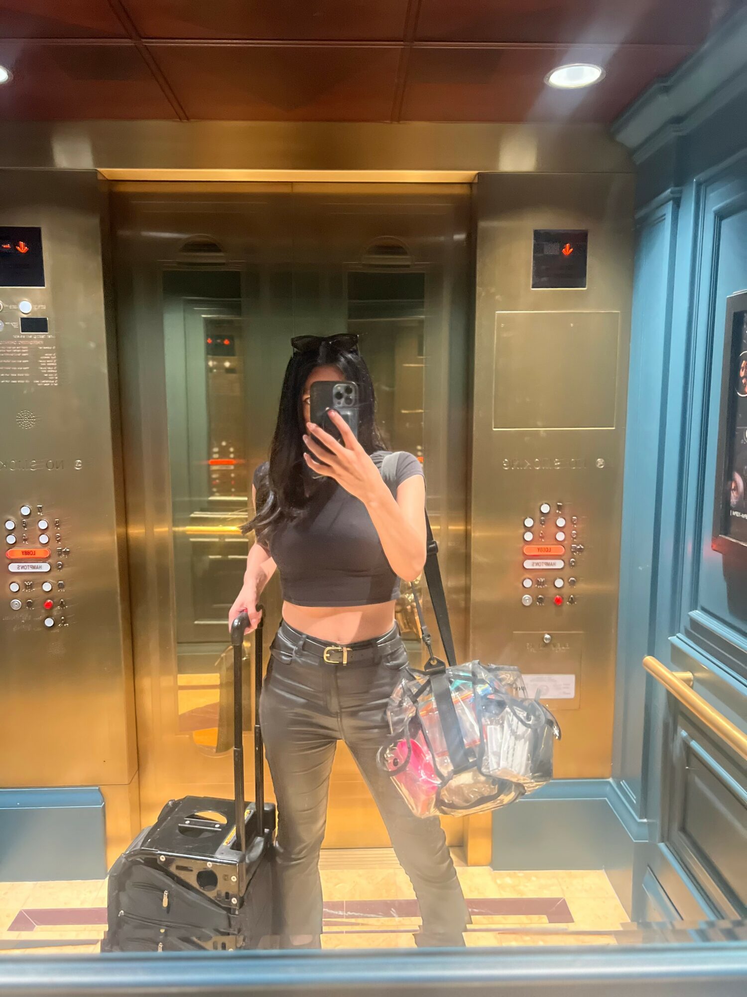 Person taking a mirror selfie in an elevator, holding a suitcase and a bag, wearing a crop top and jeans.