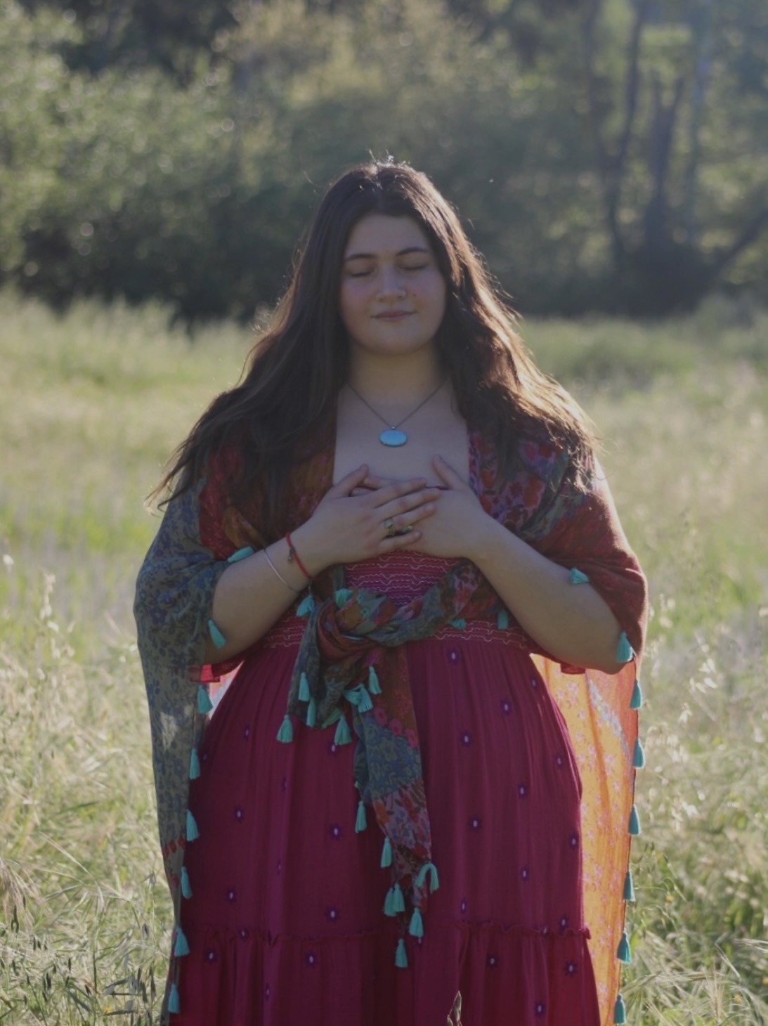Young woman standing outdoors with hands on chest, eyes closed, in a grassy field with trees in background.