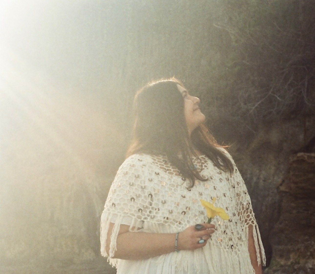 Woman with long hair holding a yellow flower, looking upward, sunlight shining from the left, outdoors with trees and rocks.