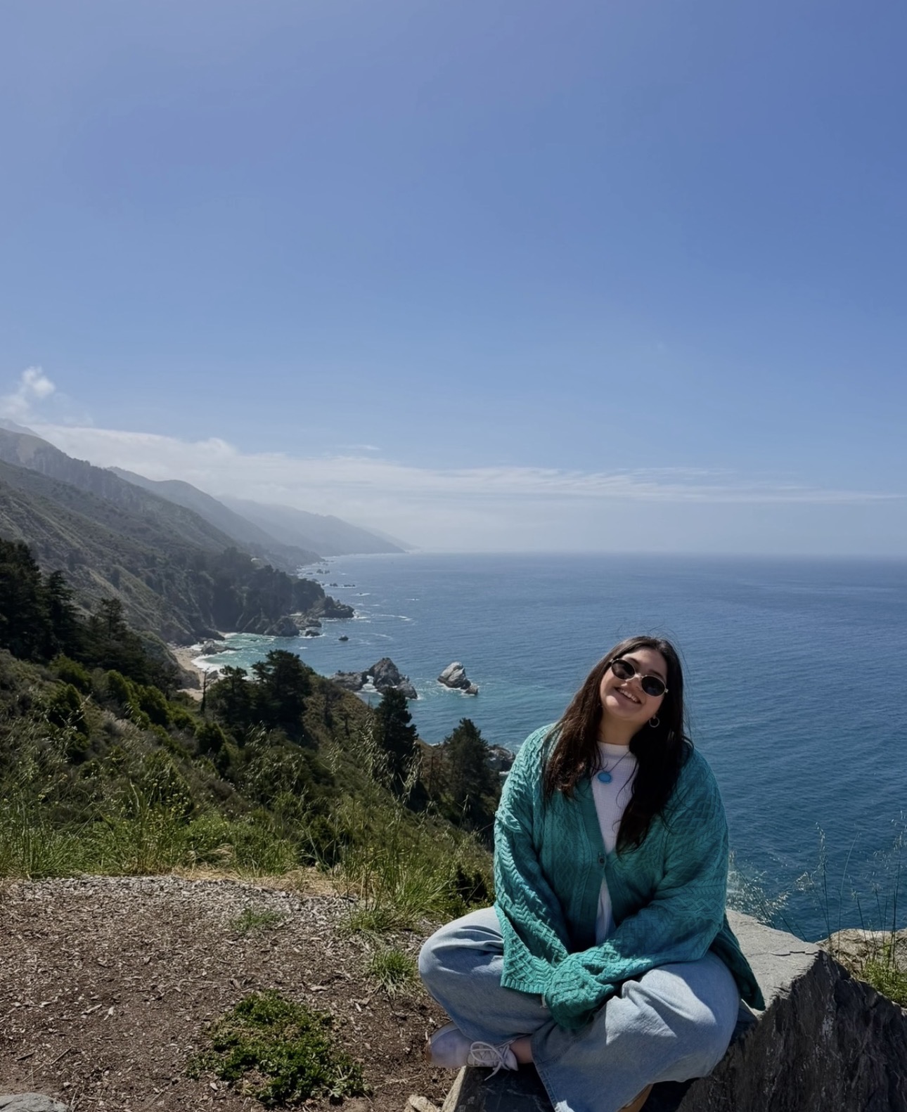 Woman sitting on a rock overlooking the ocean and coastline under a clear blue sky.
