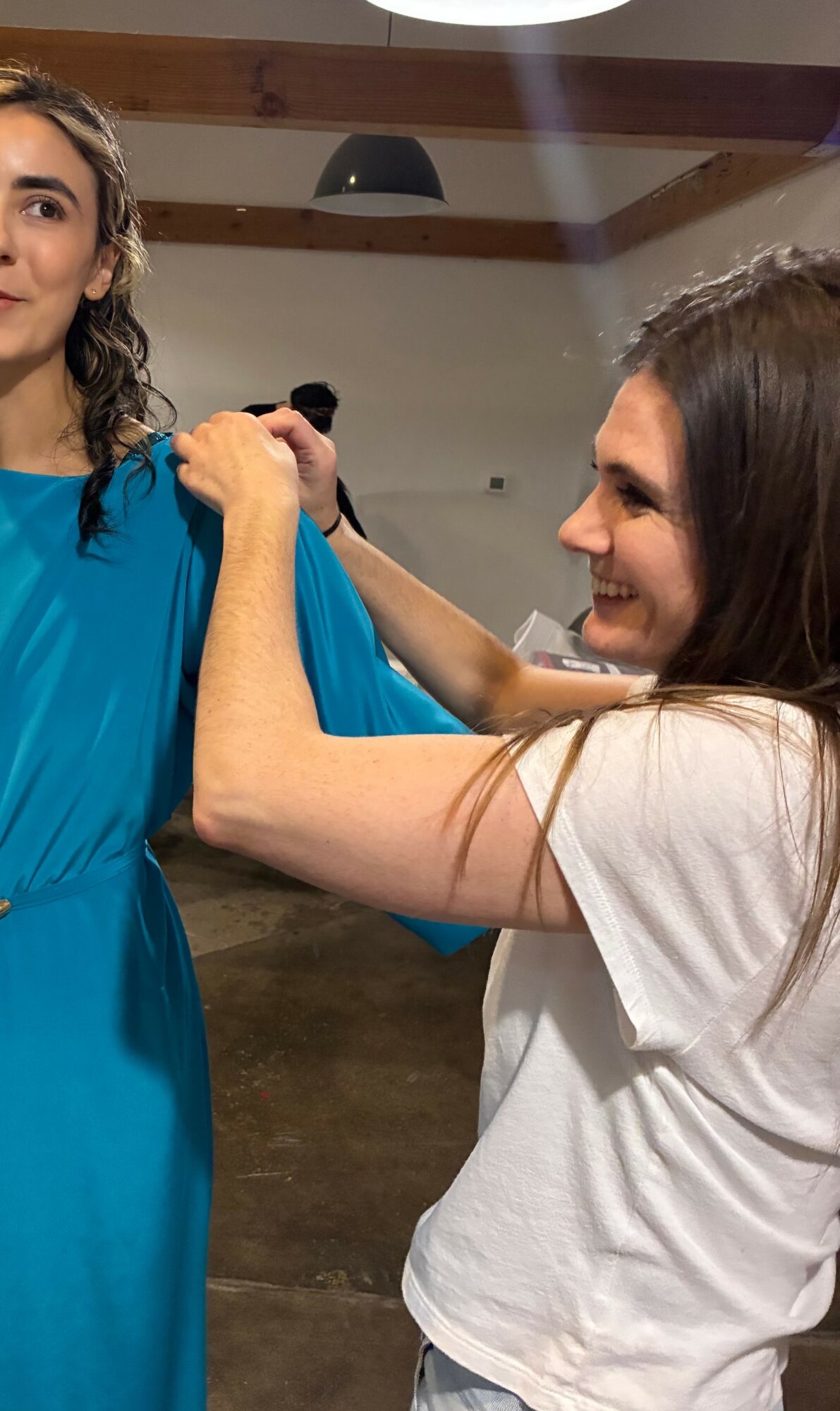 Two women smiling, one adjusting the other's blue dress, in an indoor setting with wooden beams.