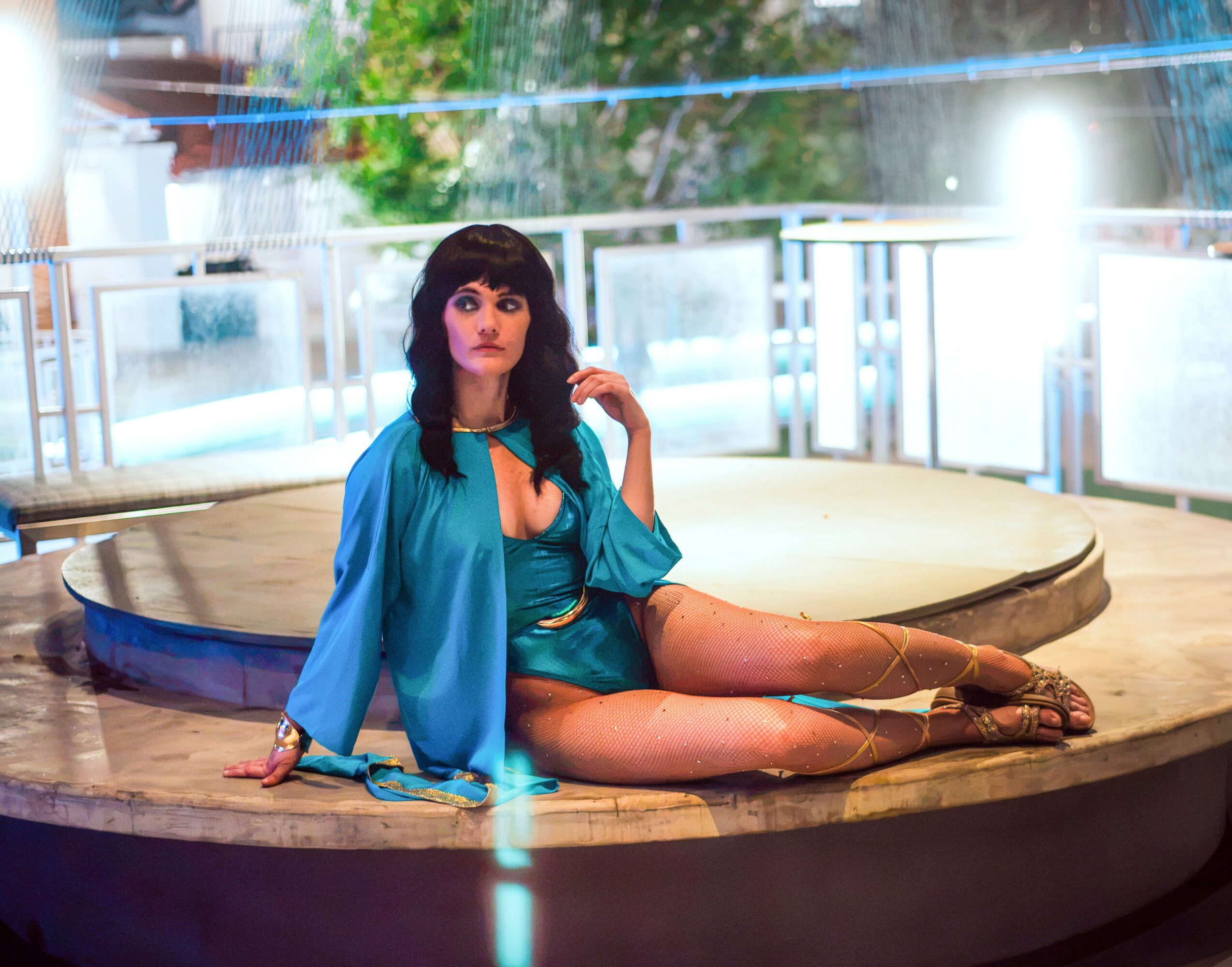 Woman with dark hair and bangs sitting on a circular stage, wearing a blue outfit, in an indoor setting.