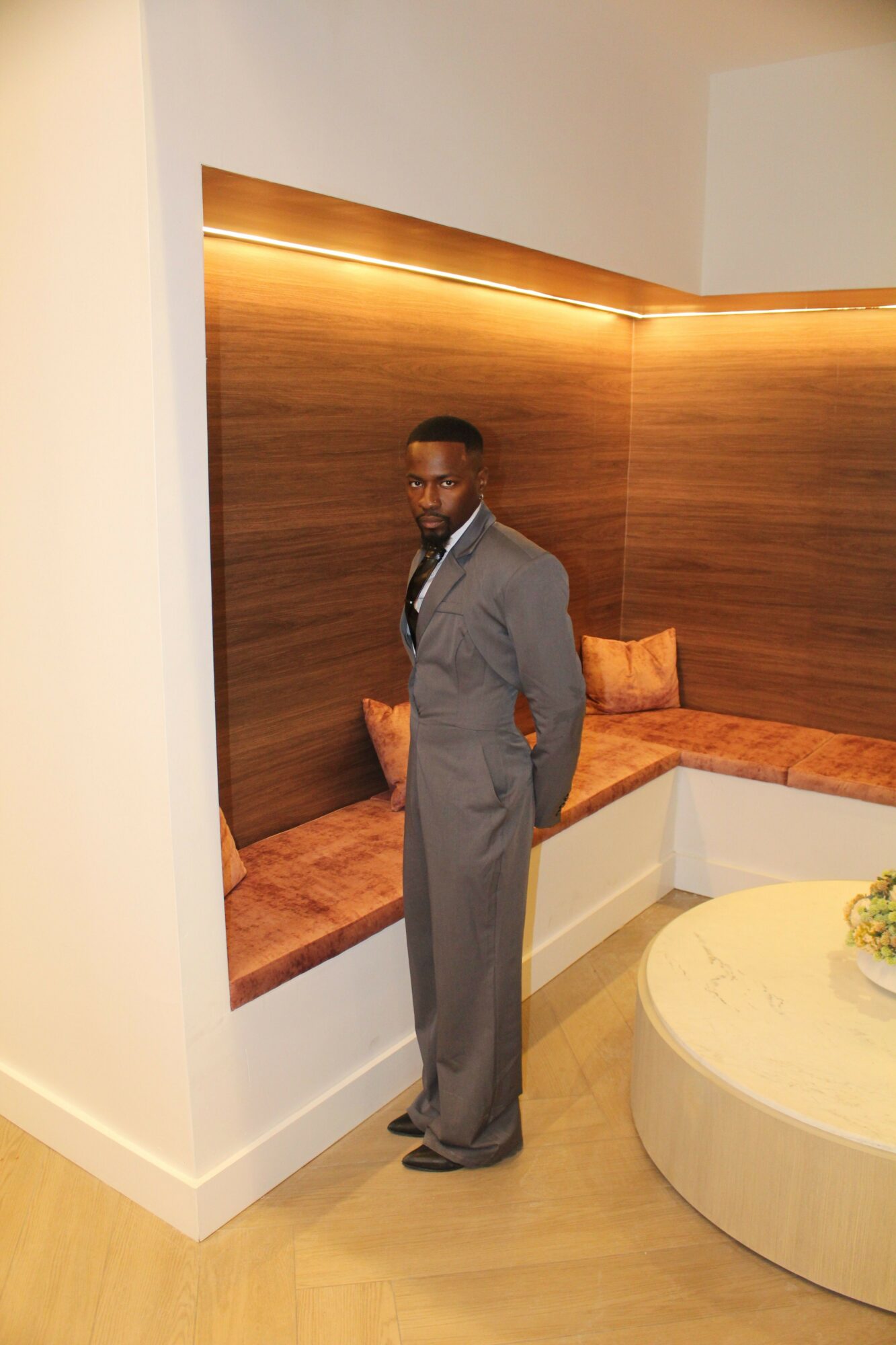Person standing in a room with wooden wall and seating area, wearing a gray jacket and dark pants.