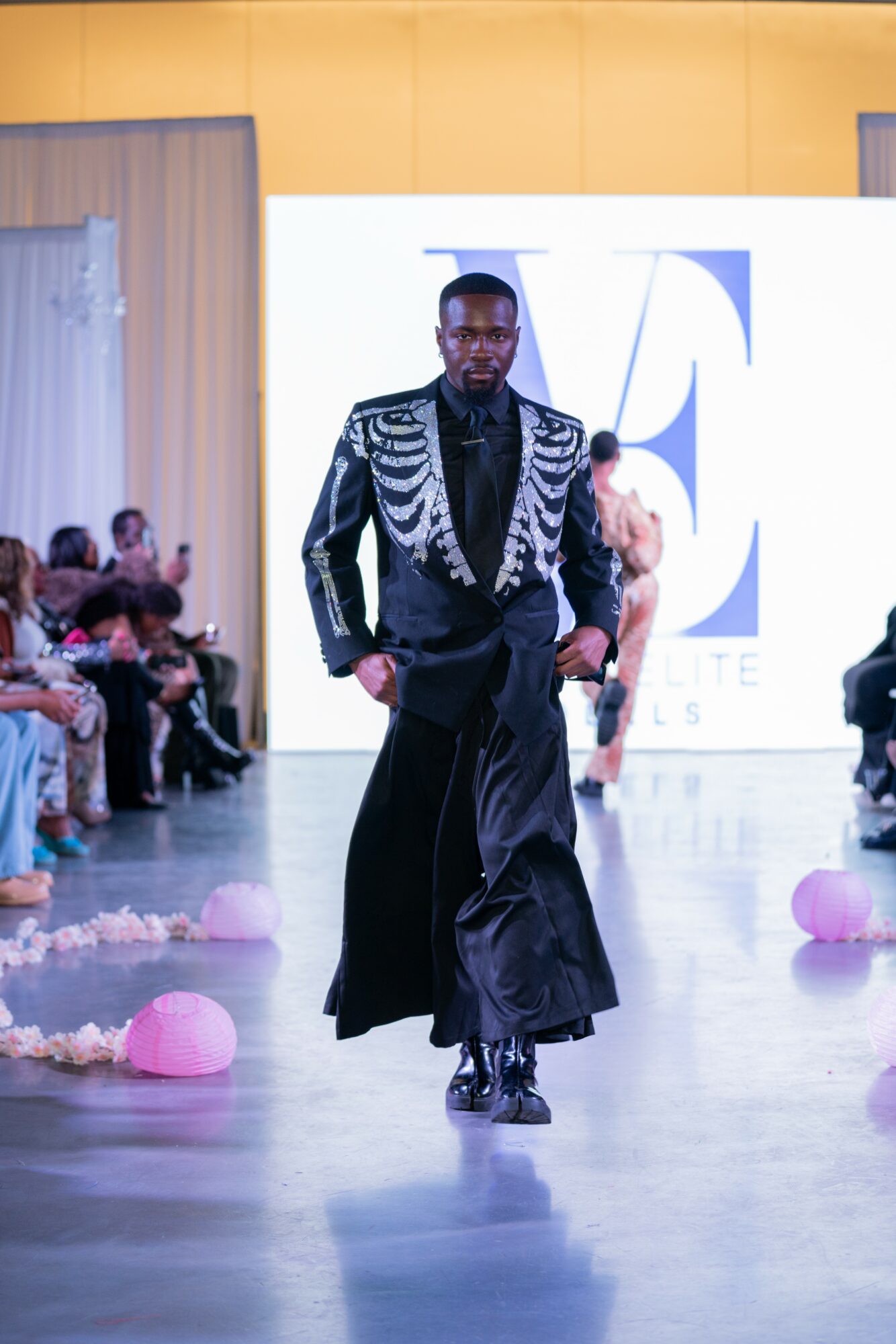 Male model walking on runway in a black and silver jacket and long black skirt, audience seated on sides, bright background.