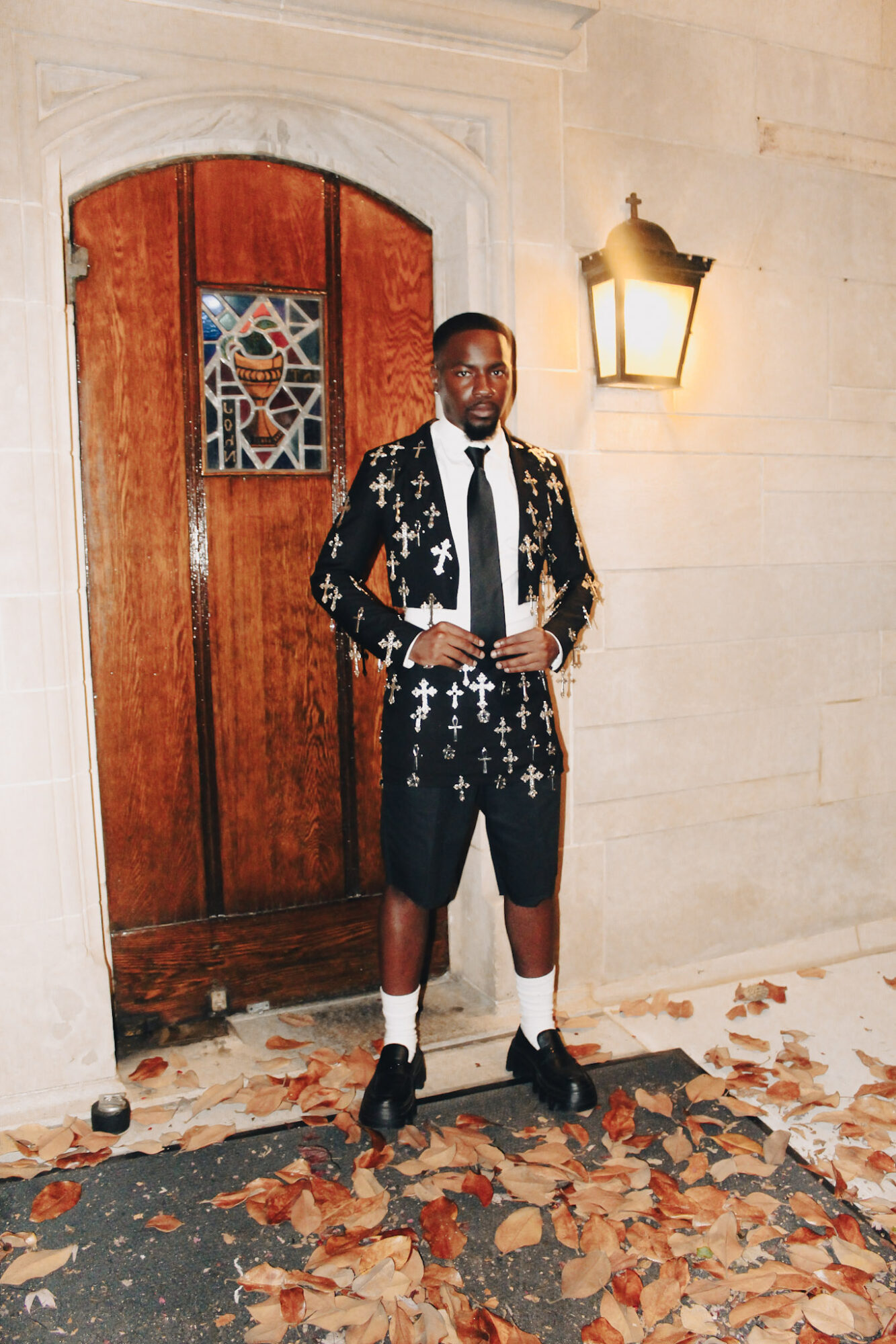 Man standing outside a wooden door with stained glass, wearing a black jacket with white crosses, black shorts, and white socks.