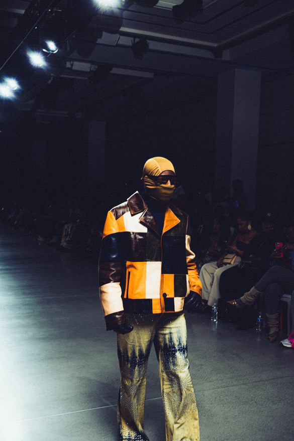Person walking on runway wearing a colorful jacket, patterned pants, and a cap, with audience in background.
