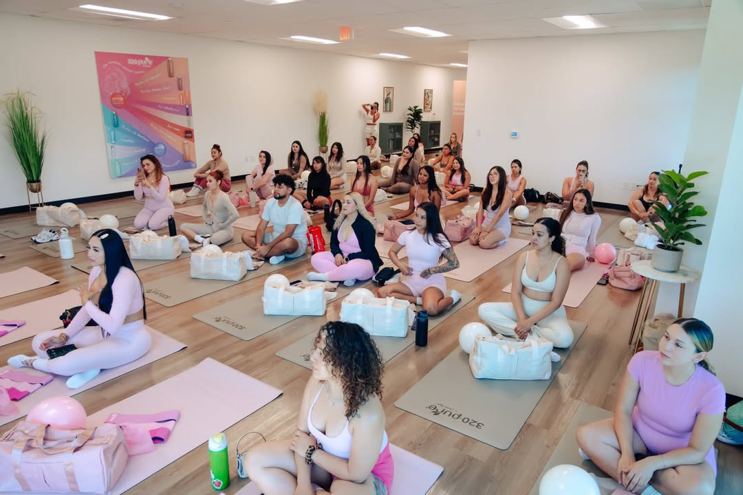 Group of people practicing yoga in a bright, spacious room with plants and colorful artwork, sitting on mats with props.