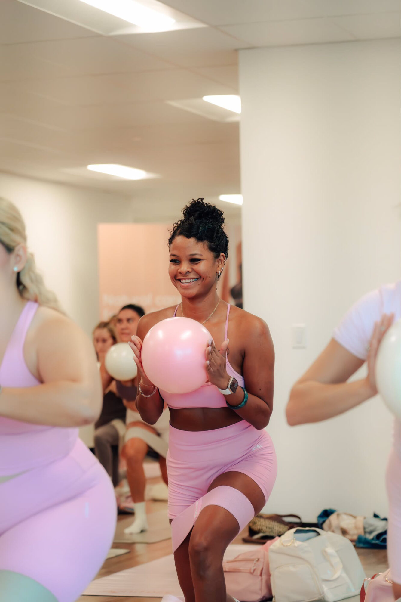 Smiling woman in pink workout outfit holding a pink ball, surrounded by others in similar attire, in an indoor setting.