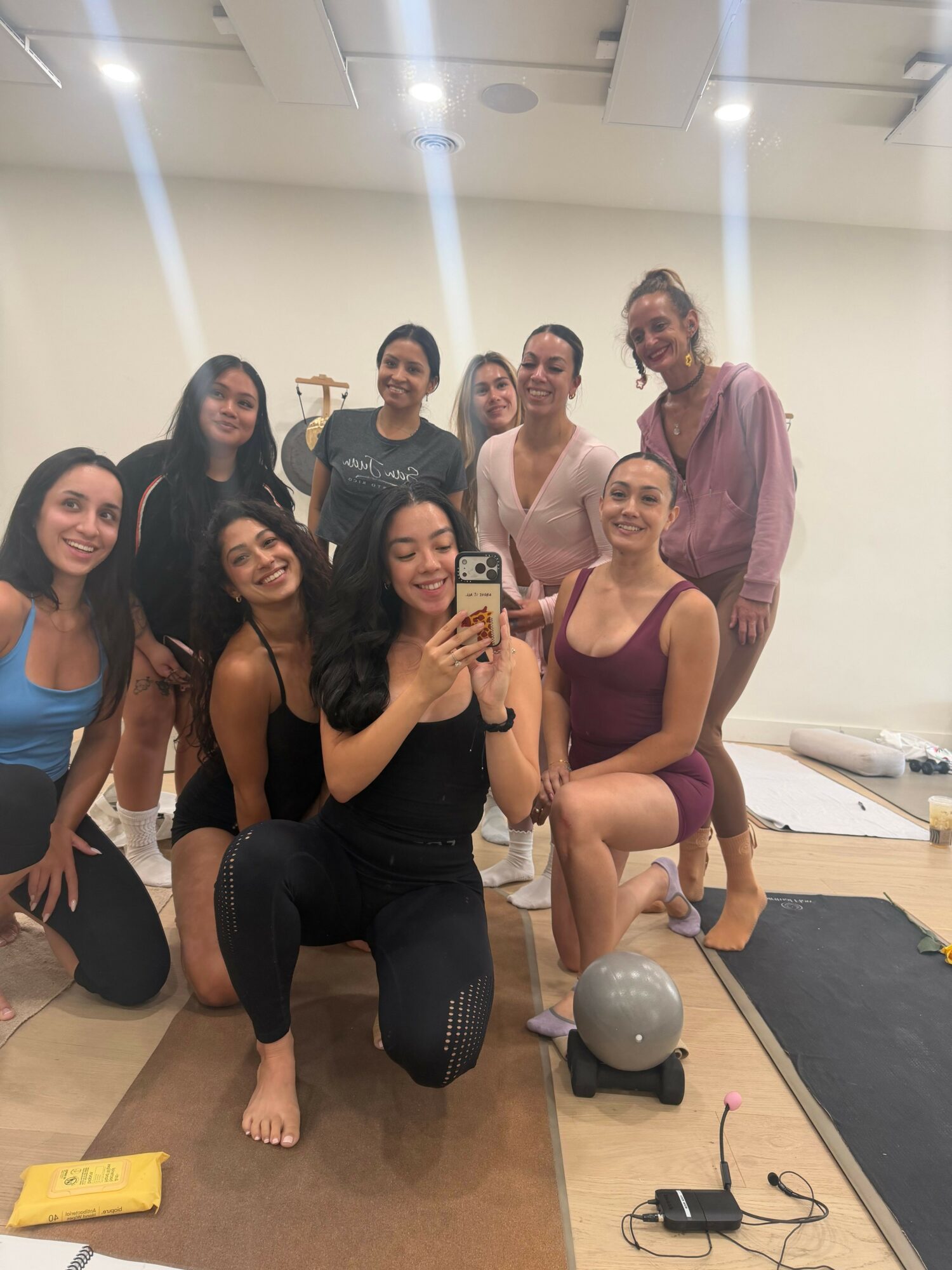 Group of seven women in workout attire taking a mirror selfie in a gym, some sitting and some standing, with gym equipment around.