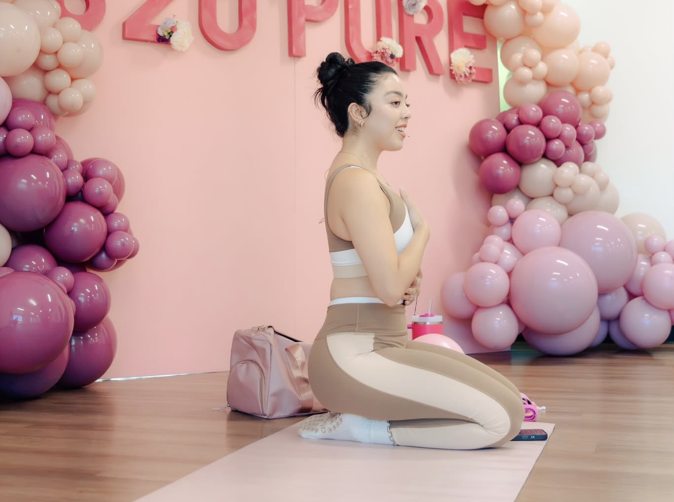 Woman practicing yoga kneeling on a mat with balloons and pink wall background, hands on chest, smiling.