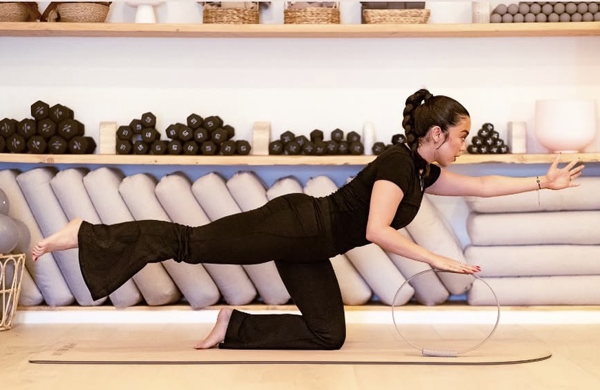 Person in black clothing performs a yoga pose on a mat in a studio with shelves of rolled yoga mats behind.