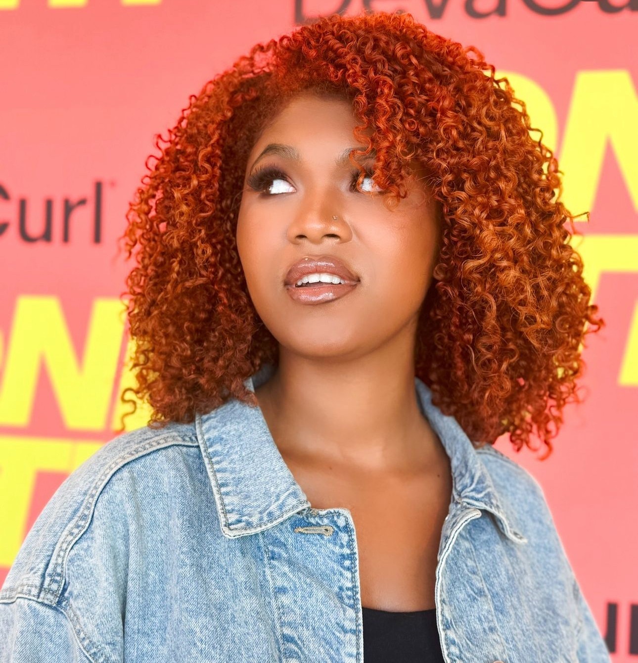 Young woman with curly red hair wearing a denim jacket, looking to the side, with a colorful background.