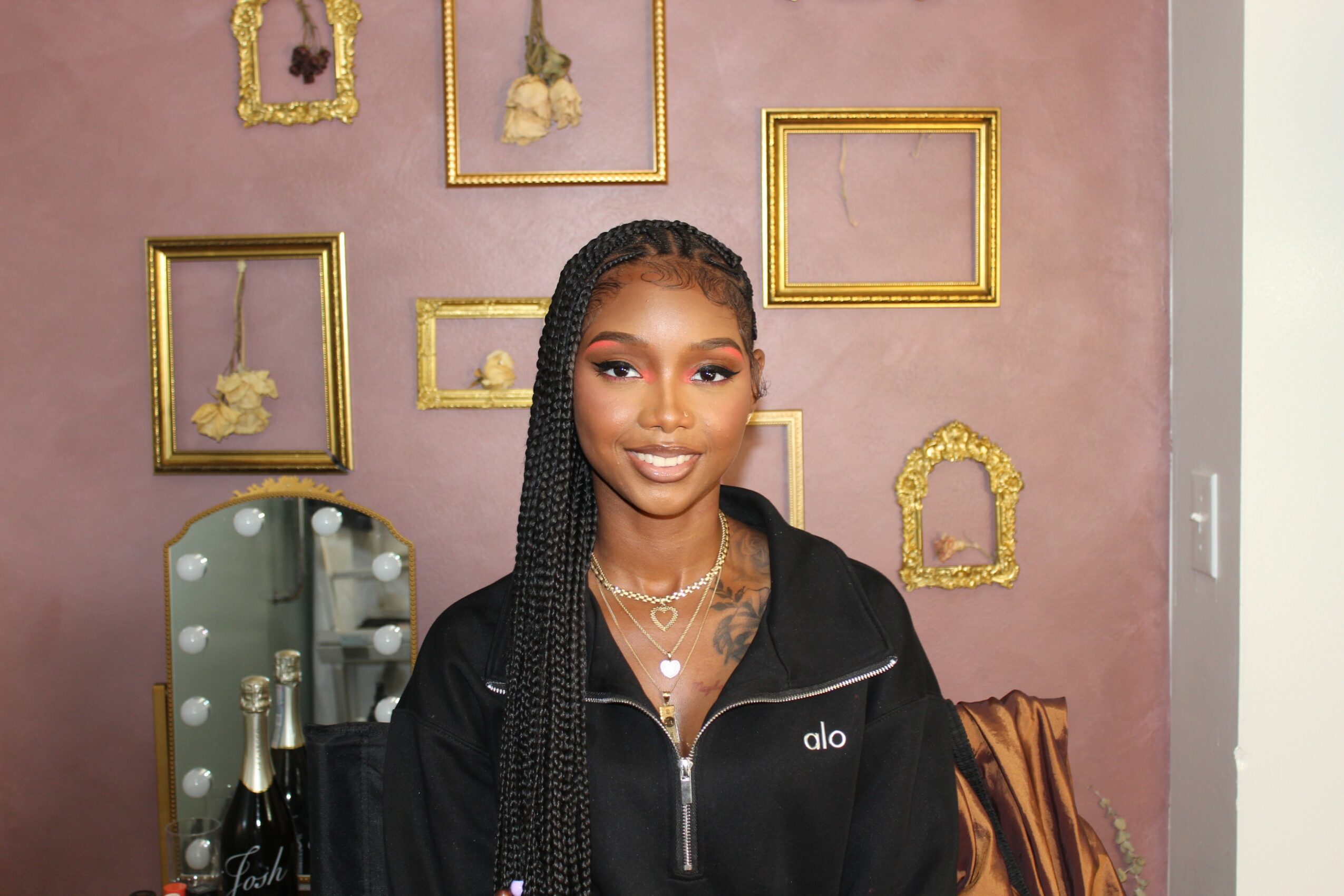 Young woman with braided hair smiling, wearing a black jacket, standing in front of a pink wall with gold picture frames.