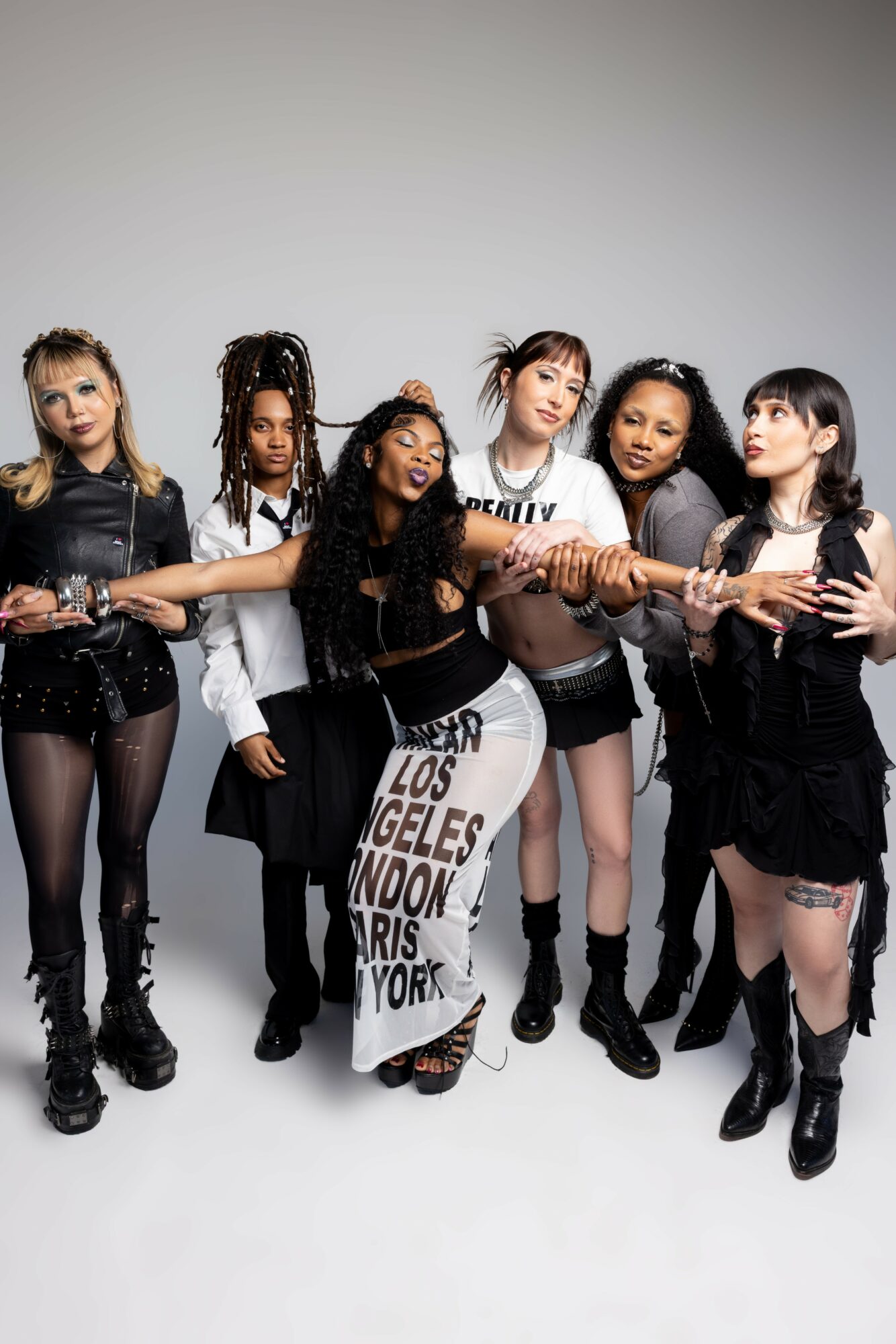 Six young women standing together holding a wooden stick, dressed in black and white outfits, in a studio setting.