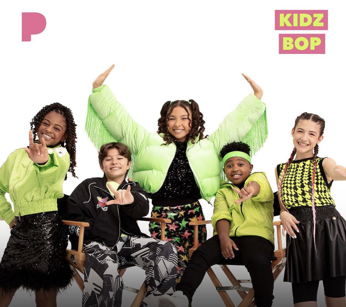Group of five children smiling, sitting and standing, wearing colorful clothing, with text 'KIDZ BOP' in the top right corner.