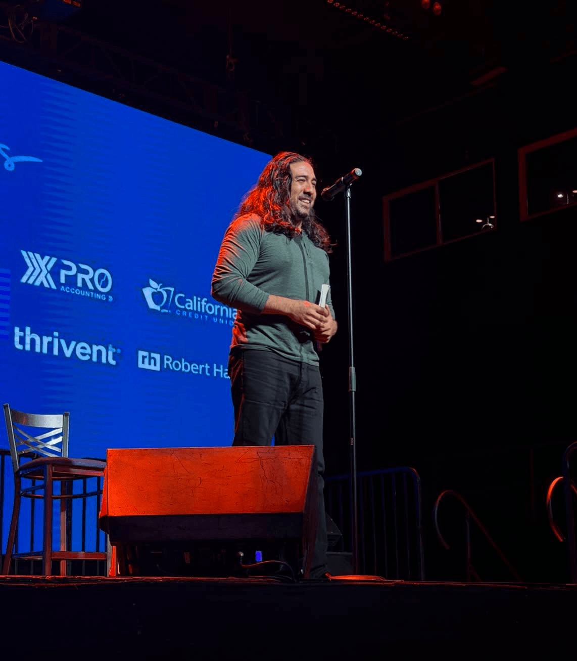 Person with long hair and beard speaking into a microphone on stage, with a blue screen behind displaying logos.
