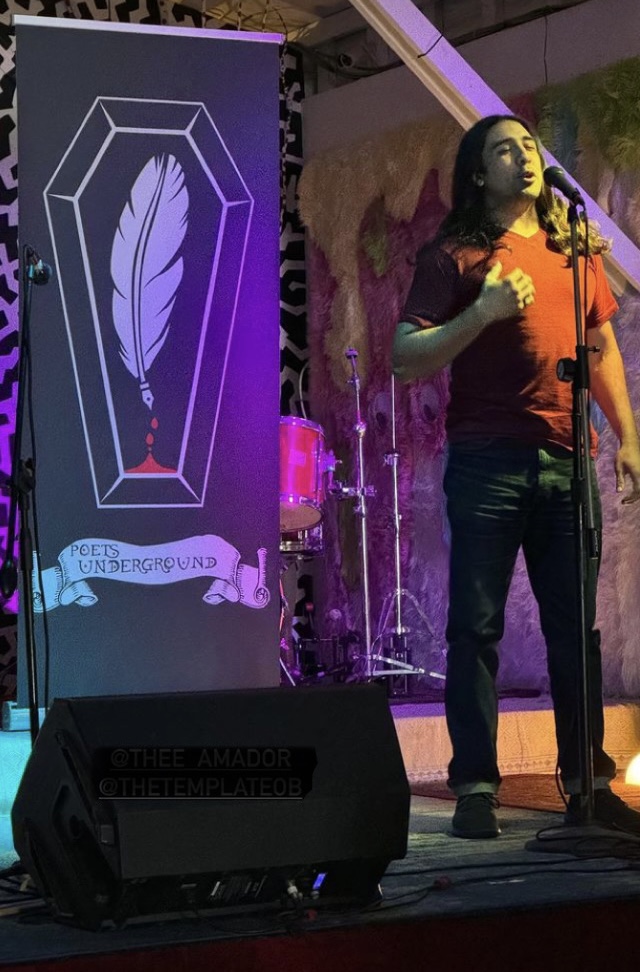 Person with long hair and orange shirt speaking into microphone on stage, purple lighting, large feather graphic on screen, musical instruments in background