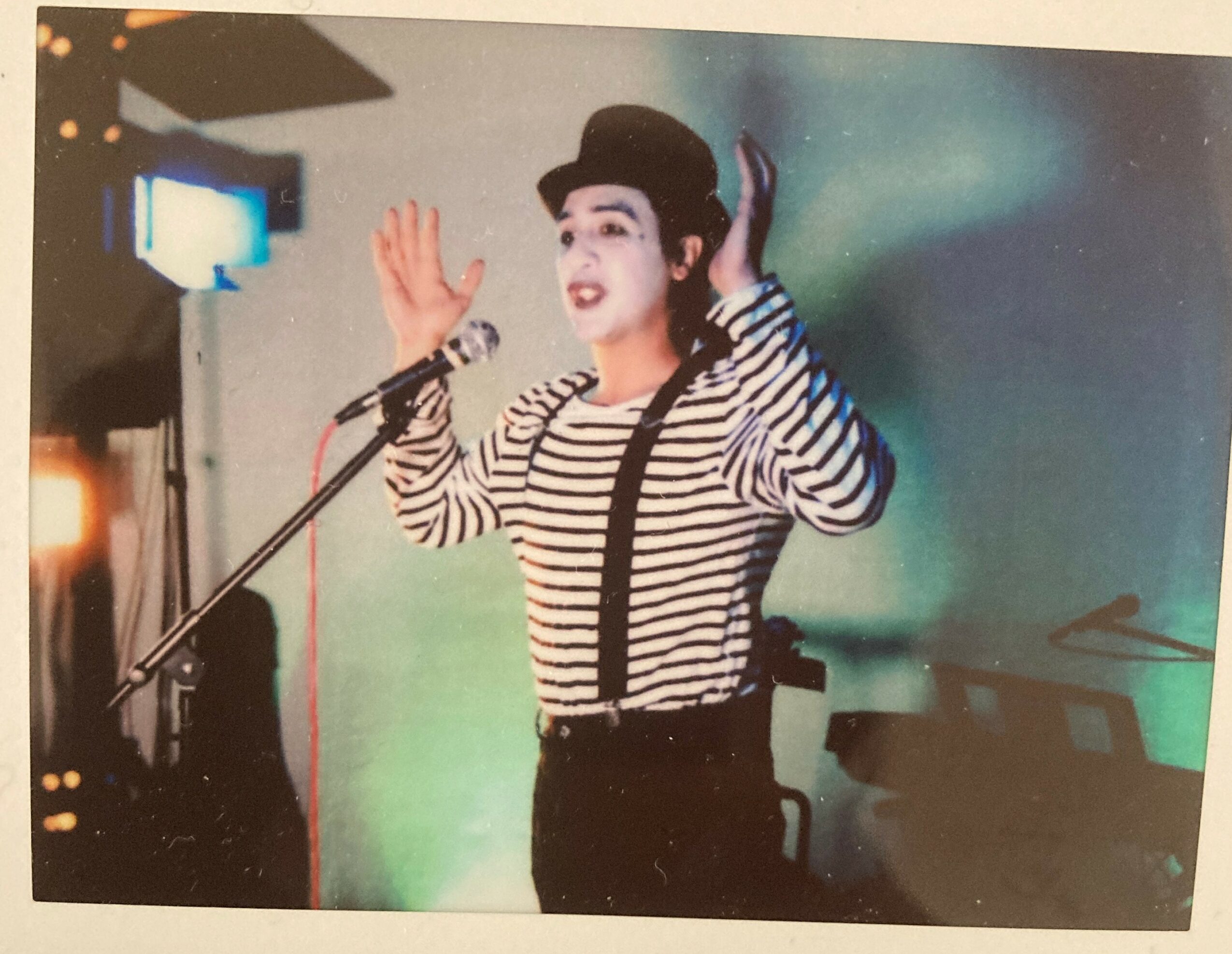 Performer with striped shirt and hat singing into microphone on stage, hands raised, with piano in background.