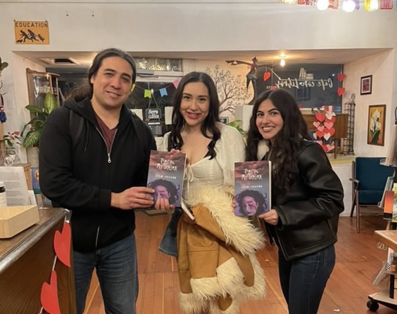 Three people standing indoors, holding books, smiling, with a decorated background and warm lighting.