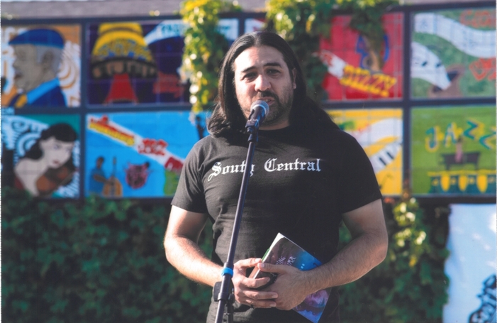 Person with long dark hair speaking into a microphone outdoors, holding a book, with colorful mural in background.