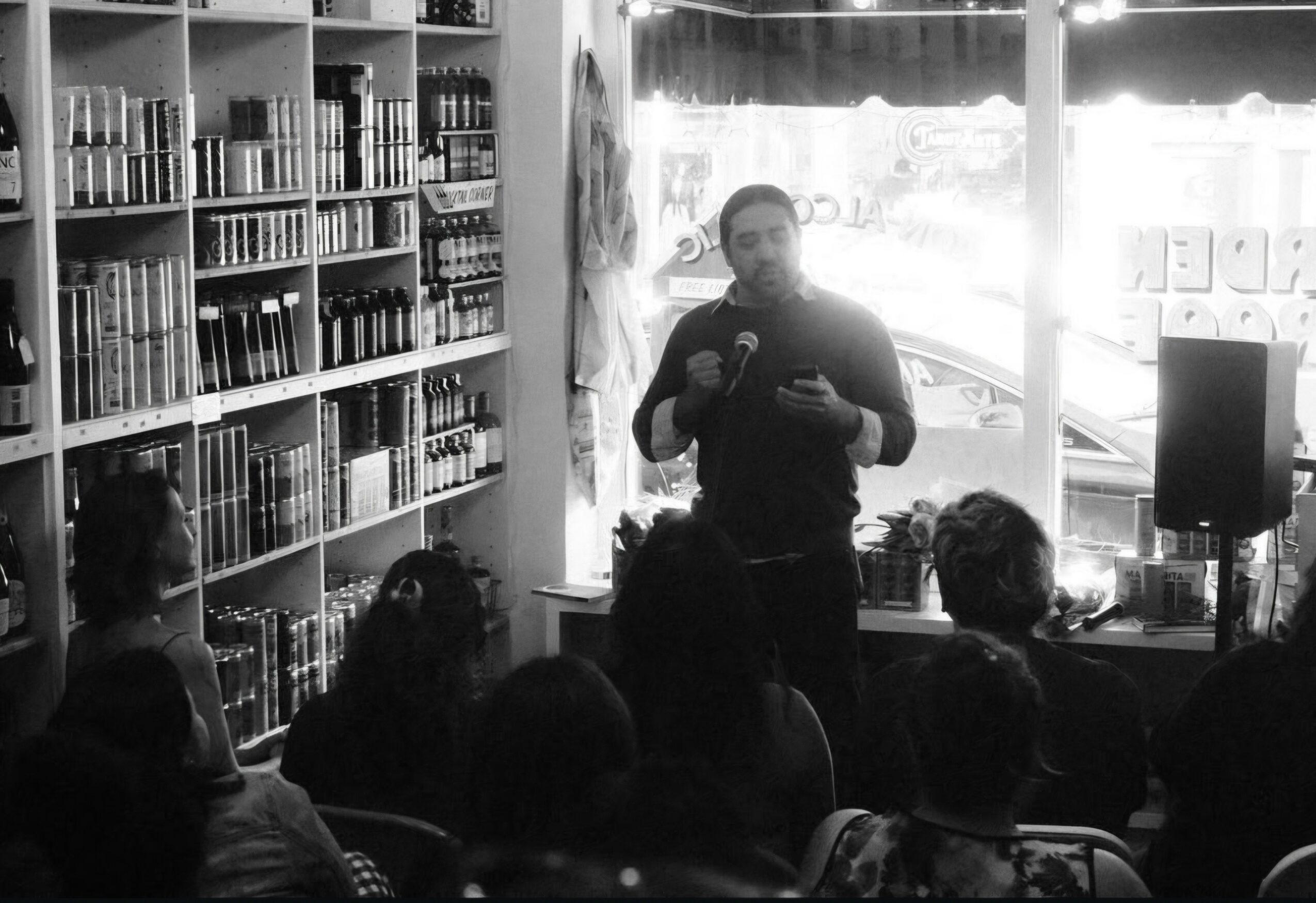 Person speaking to an audience in a bookstore, shelves filled with books behind, sunlight through window, audience seated facing speaker.