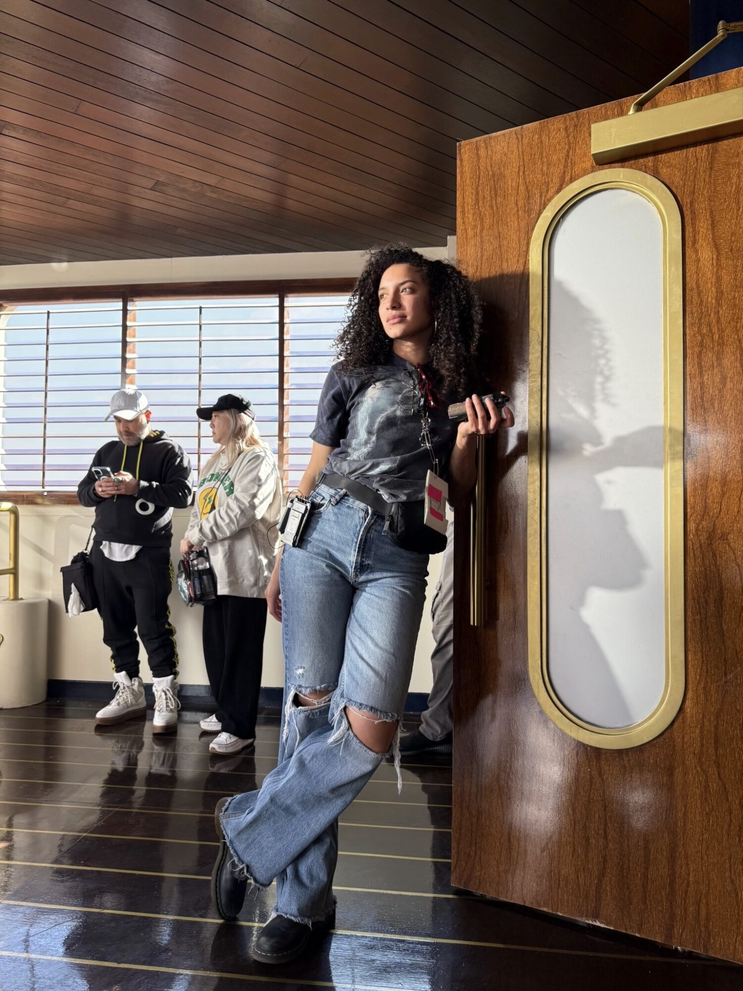 Young woman with curly hair leaning against a wooden wall in a modern interior, with people in the background, wearing casual clothing.