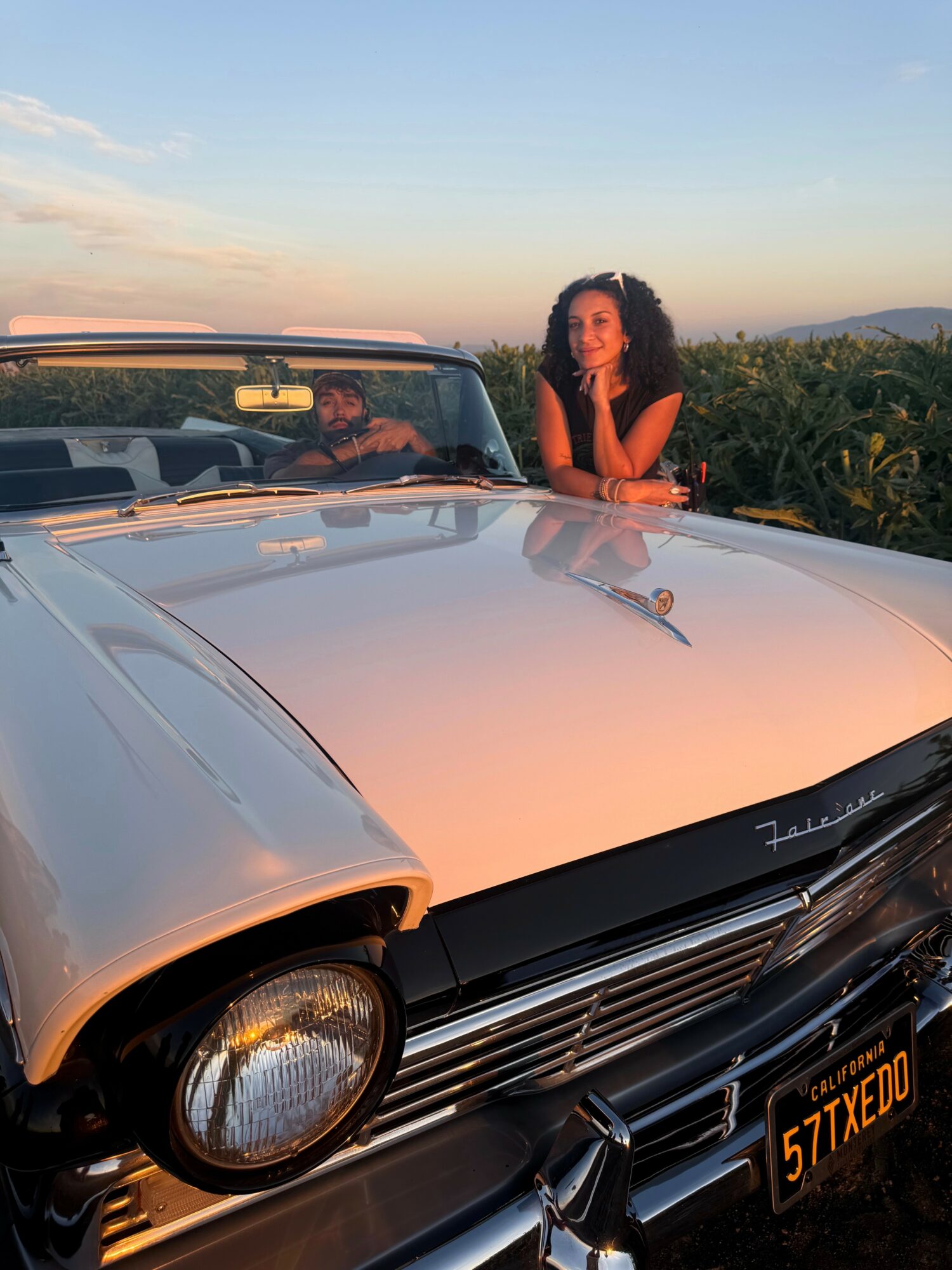 Woman with curly hair leaning on a vintage car's hood, with a person in the background driving, outdoors during sunset.