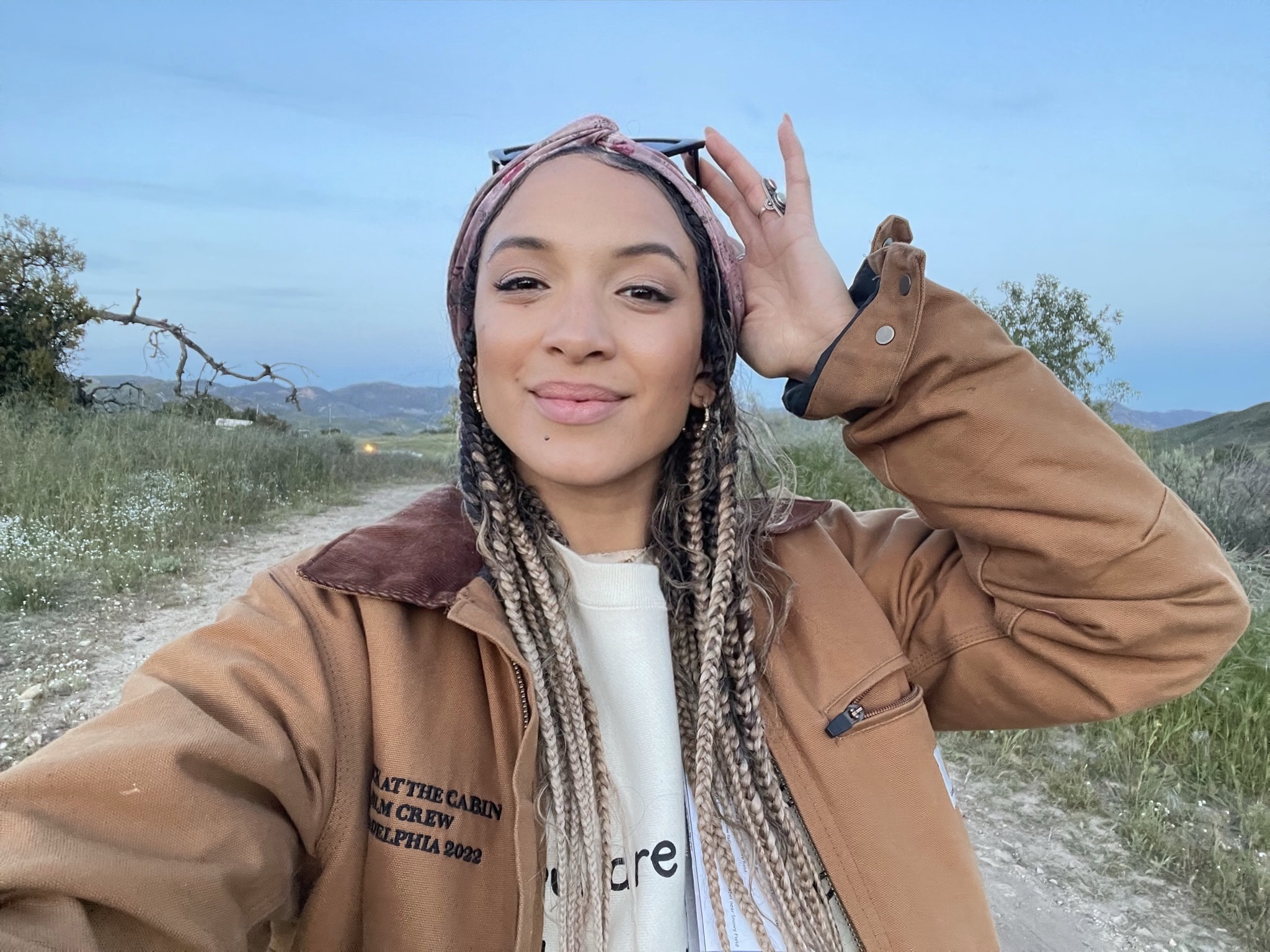 Young woman outdoors with braided hair, smiling, wearing a brown jacket and headband, on a dirt path with greenery and blue sky.