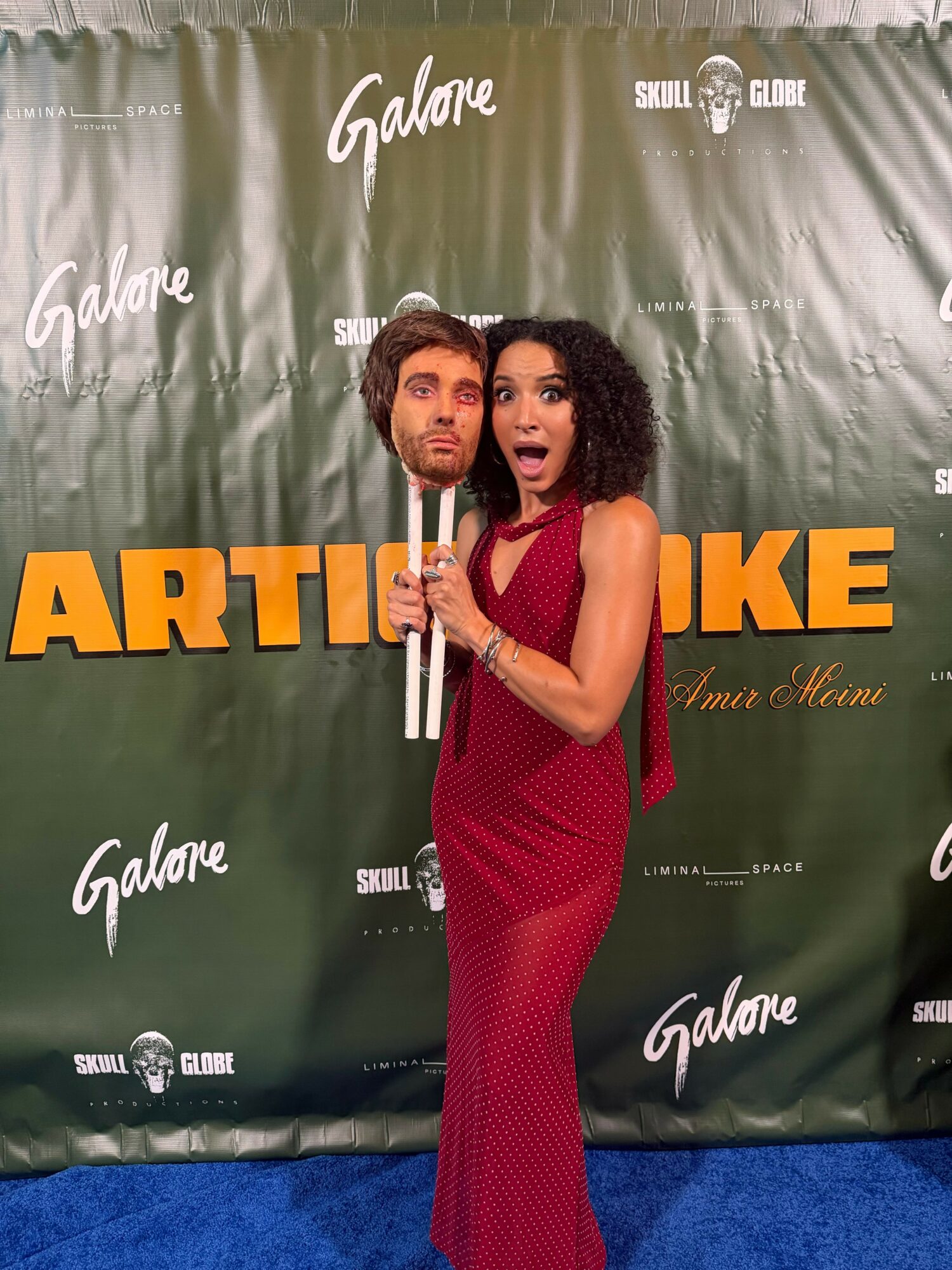 Woman in a red dress holding a mask with a man's face, standing on a blue carpet in front of a green backdrop with logos.