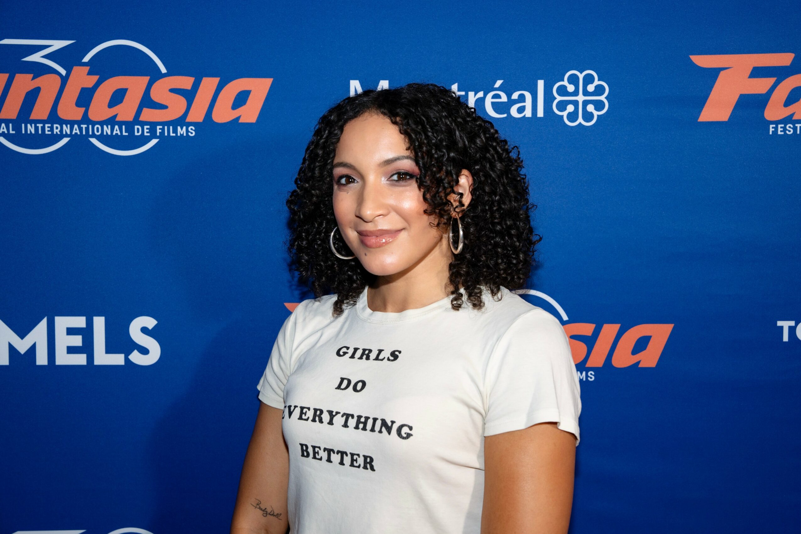 Young woman with curly hair smiling in front of a blue background with logos, wearing a white T-shirt with text.