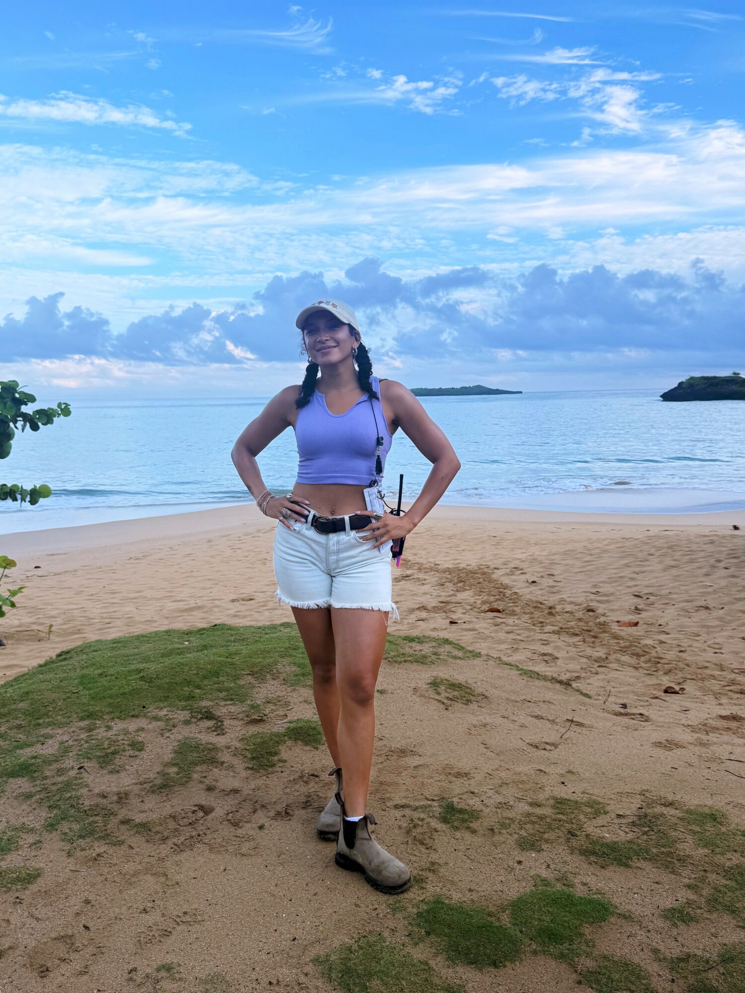 Young woman standing on a beach with water and sky in background, smiling, wearing a purple top, white shorts, and boots.